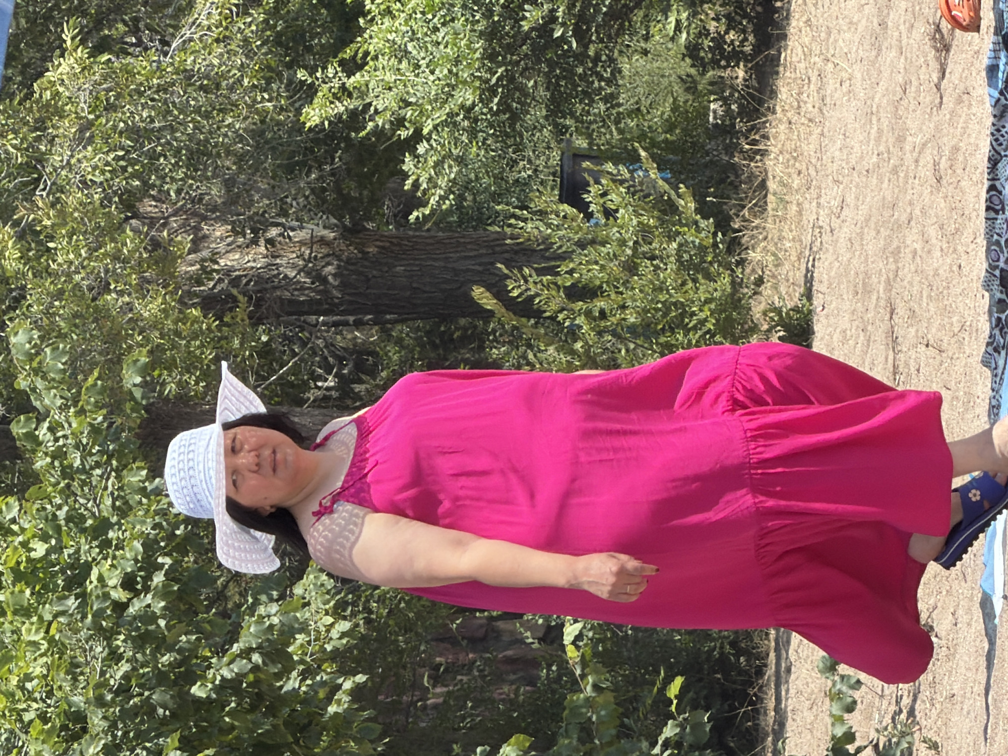 A medium shot captures a woman standing in a sandy area, partially shaded by trees. She wears a wide-brimmed white hat and a flowing, bright pink maxi dress. Her left arm is bent at the elbow, resting near her side, while her right arm is not fully visible. She appears to be looking towards the camera. In the background, lush green foliage and trees create a natural setting. The ground is covered in sand and dry grass. A patterned cloth or blanket is laid out on the sand in the foreground, with one of the woman's feet resting on it. She is wearing blue sandals with a floral embellishment. The lighting suggests it is daytime, with sunlight filtering through the leaves.