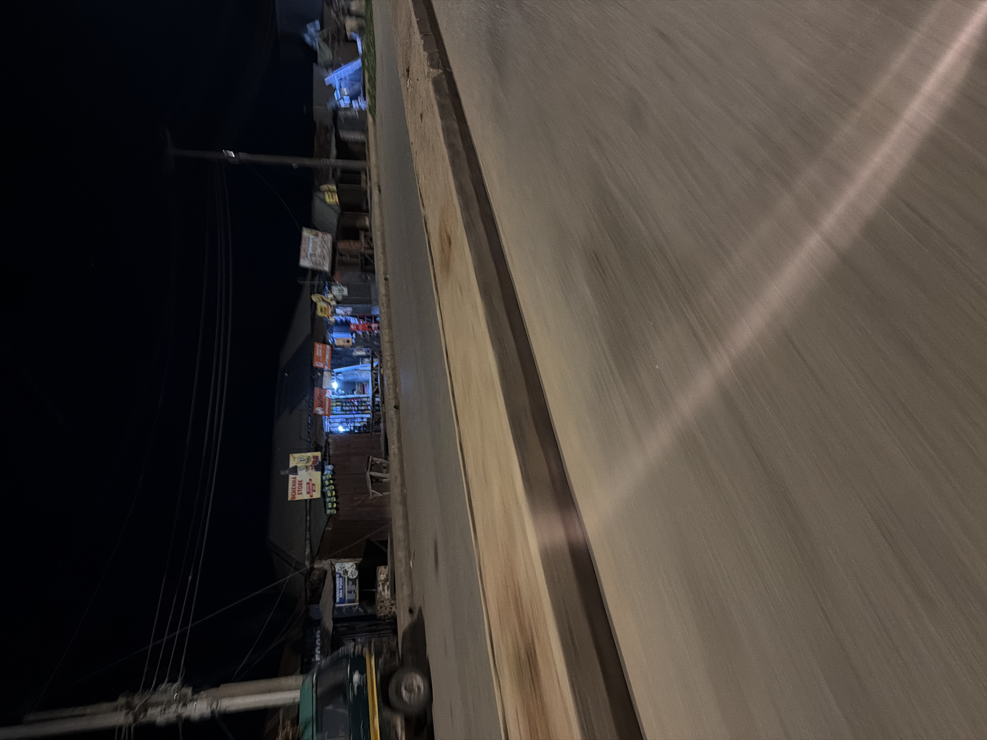 The image depicts a street scene at night in Bukuru, Nigeria. The perspective is from a moving vehicle, indicated by the motion blur on the road. The left side of the frame shows a dark area with utility poles and wires. Adjacent to this is a row of shops or stalls, illuminated by artificial light, displaying various goods. Some signs are visible, including one that reads "OKENCHA STORE." The shops appear to be open for business, with merchandise visible on shelves and in displays. There are no people clearly visible, but the presence of active businesses suggests nighttime commerce. The road itself is paved and shows a distinct lane marking or debris line. A light beam, likely from a streetlamp or another vehicle, cuts diagonally across the road surface, creating a bright streak. The overall atmosphere is one of a lively, albeit dimly lit, commercial street after dark.
