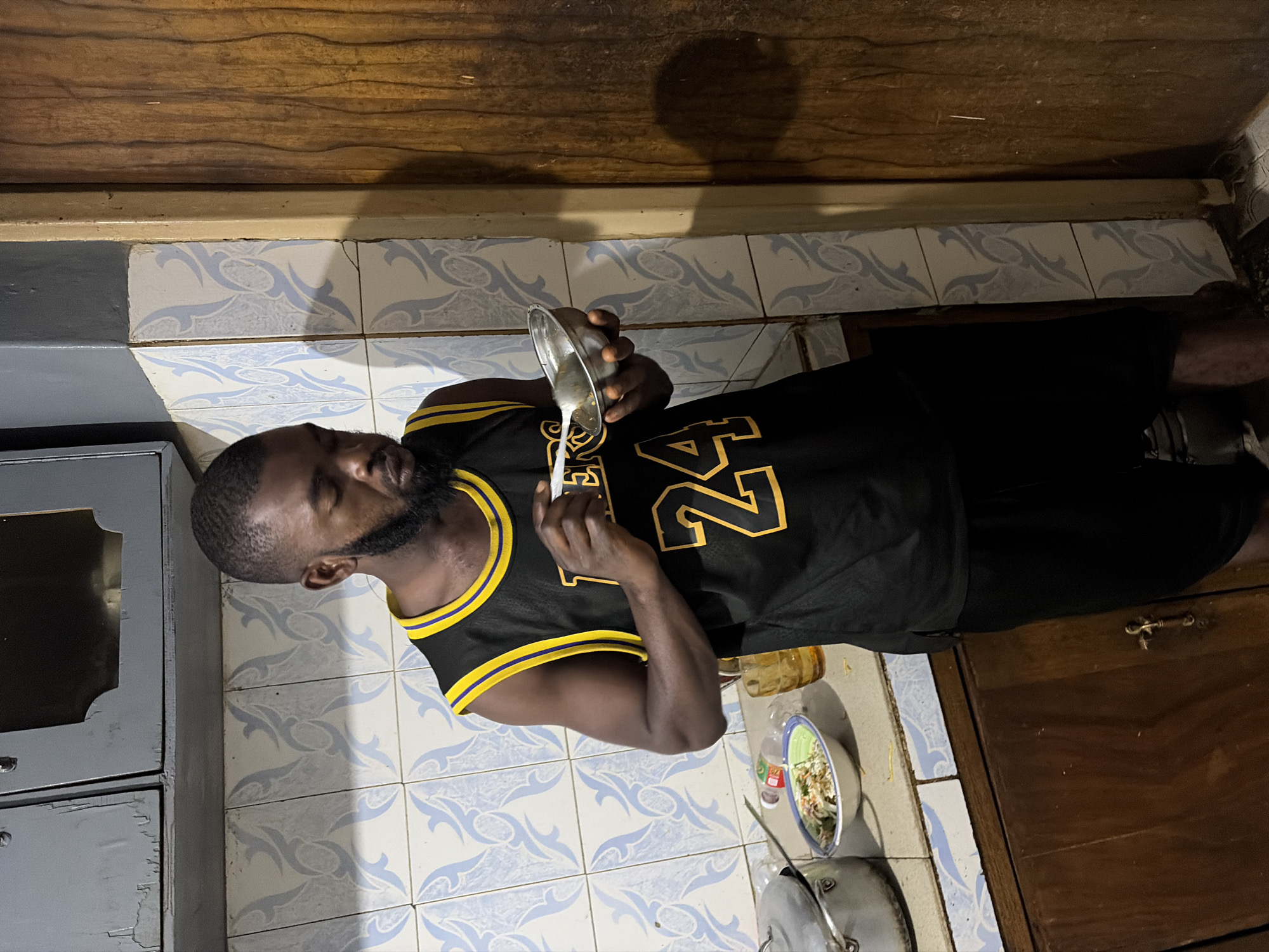 A Black man, appearing to be in his late 20s or early 30s, stands in what appears to be a kitchen environment. He is wearing a black basketball jersey with yellow trim and the number "24" in yellow on the front. He has a short beard and is looking down at a small metal bowl he is holding in his left hand, which contains some food. He is using a white spoon in his right hand to eat from the bowl.
The kitchen has light blue and white patterned tiles on the wall behind him, and grey cabinets above the counter. On the counter to his left, there is a metal pot, a bowl of salad, and a glass with a yellow liquid. To his right, there is a wooden door. The overall setting suggests a modest, possibly older, kitchen. The image was taken in Jos, Nigeria.