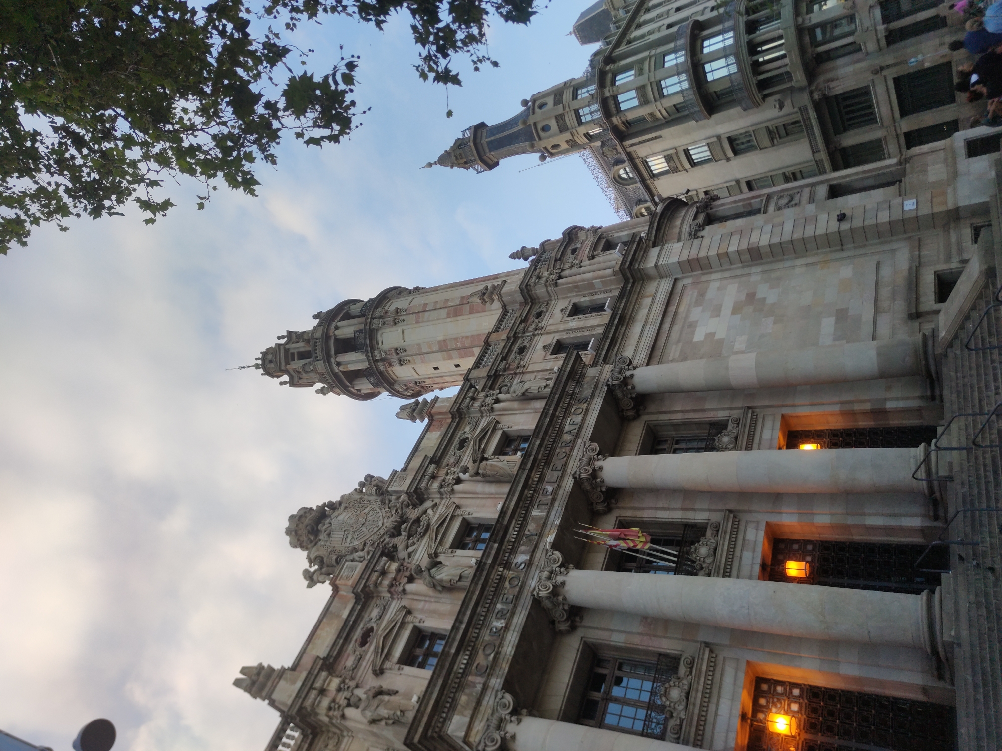 The image shows a low-angle view of the Barcelona Post Office and Telegraph Building (Palau de Correos i Telègrafs) in Barcelona, Spain, on a partly cloudy day. The grand Beaux-Arts building features ornate stone carvings, large columns, and a prominent tower. To the right of the main building, another building with a distinct pointed tower is visible. A tree with green leaves extends from the top right corner of the frame, partially obscuring the sky. The ground level of the main building has large, dark doors and is lit by warm yellow lamps. A set of stone stairs leads up to the entrance. In the foreground, at the bottom right, a small group of people, mostly with their backs to the camera, are standing on the sidewalk. One person is wearing a bright pink top. The building inscription "CORREOS Y TELEGRAFOS" is visible above the columns.