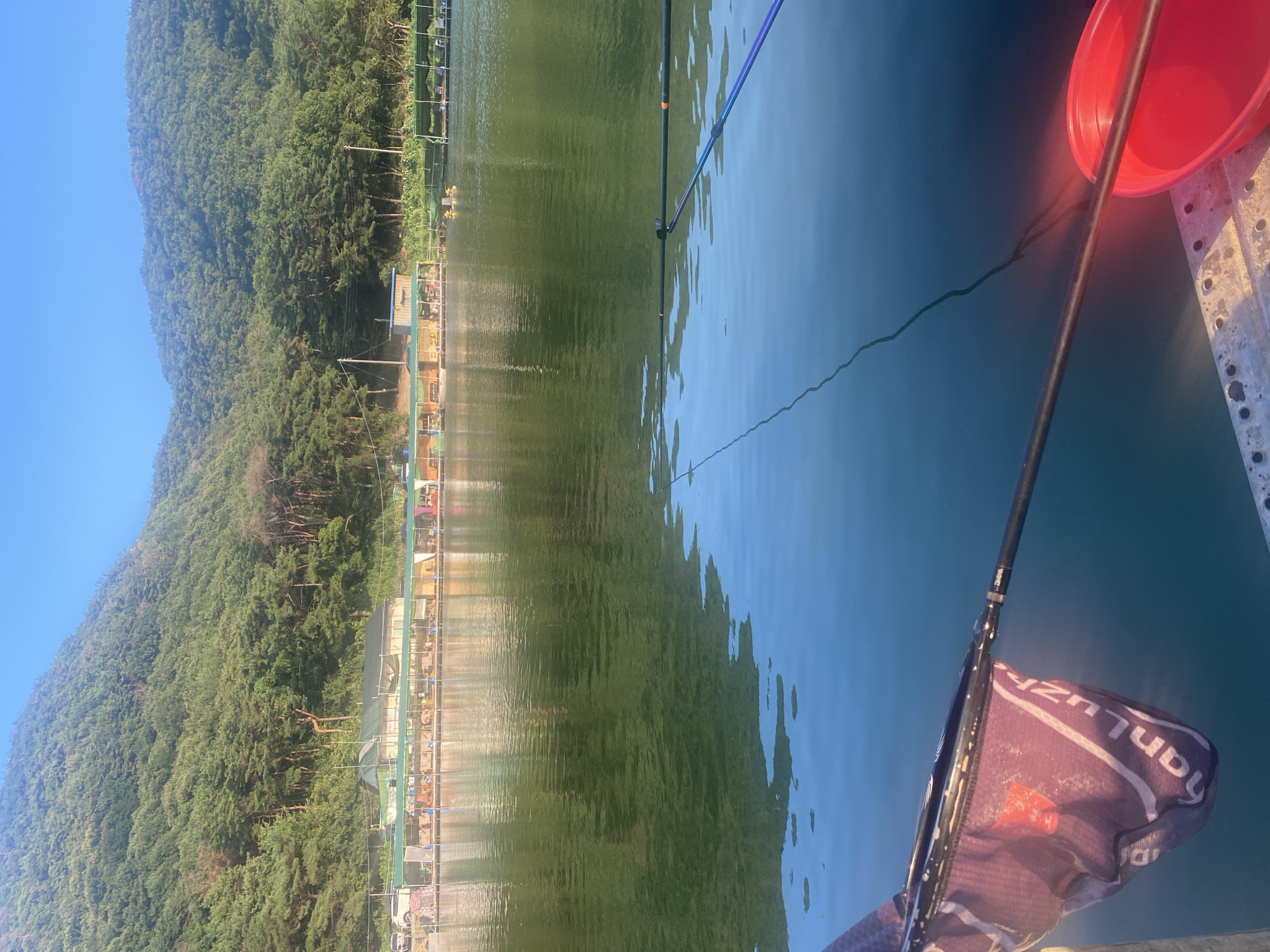 A serene waterscape captures a fishing setup on a calm lake. The foreground is dominated by fishing gear, including a purple net with visible text reading "nanLuzh" and a bright red bucket, positioned on a metallic platform. Two fishing rods extend into the water, their lines disappearing into the still surface, reflecting the verdant, tree-covered mountains that rise majestically in the background. Along the shoreline, a row of covered fishing platforms, occupied by a few anglers, creates a sense of leisurely activity. The clear blue sky suggests a pleasant day for fishing. This scene is likely at a fishing resort or recreational area, possibly on the Nakdong River or one of its associated lakes in Yangsan-si, South Korea, known for its natural beauty and outdoor activities.