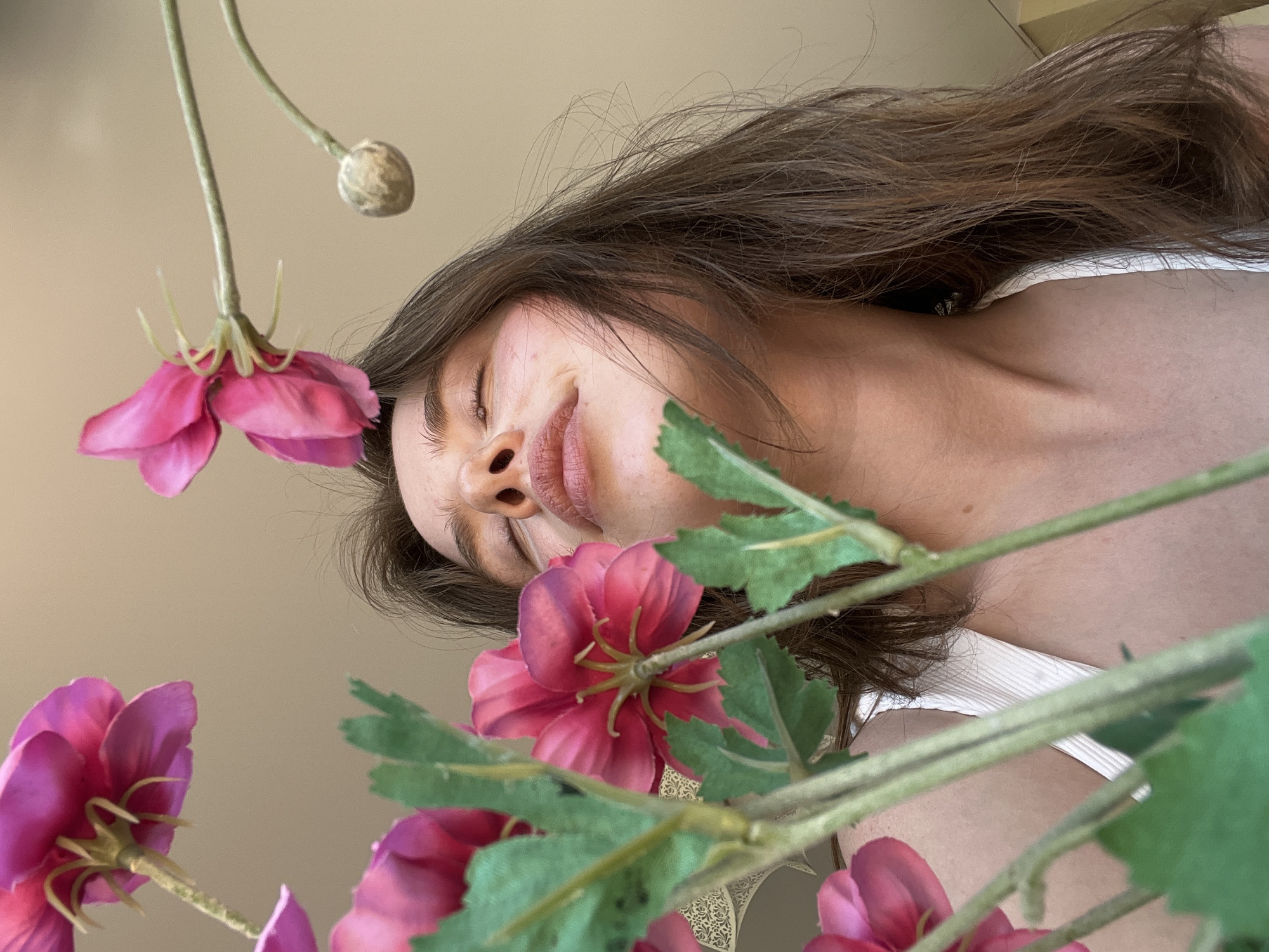 The image captures a young woman from an extreme low-angle perspective in an indoor setting. Her eyes are closed, and her head is tilted back, positioning her face amidst several artificial pink flowers, suggesting an action of smelling or serene contemplation. Her facial features include a prominent nose with visible nostrils, slightly parted lips, and dark, full eyebrows. Long, dark brown hair frames her face and extends around her shoulders. She wears a light-colored, ribbed tank top, visible on her upper chest and shoulder.
The artificial flowers, characterized by vibrant pink petals and green stems with visible leaves, are arranged in the foreground and upper sections of the frame. One flower is positioned directly below her nose, near her mouth. Another closed bud is visible in the upper right. A faint, light-colored embossed pattern is partially discernible on a surface in the background on the left side. The overall background is a plain, uniform light beige or tan, consistent with an interior wall or ceiling. This scene is situated in the city of Chaikovsky, Russia.