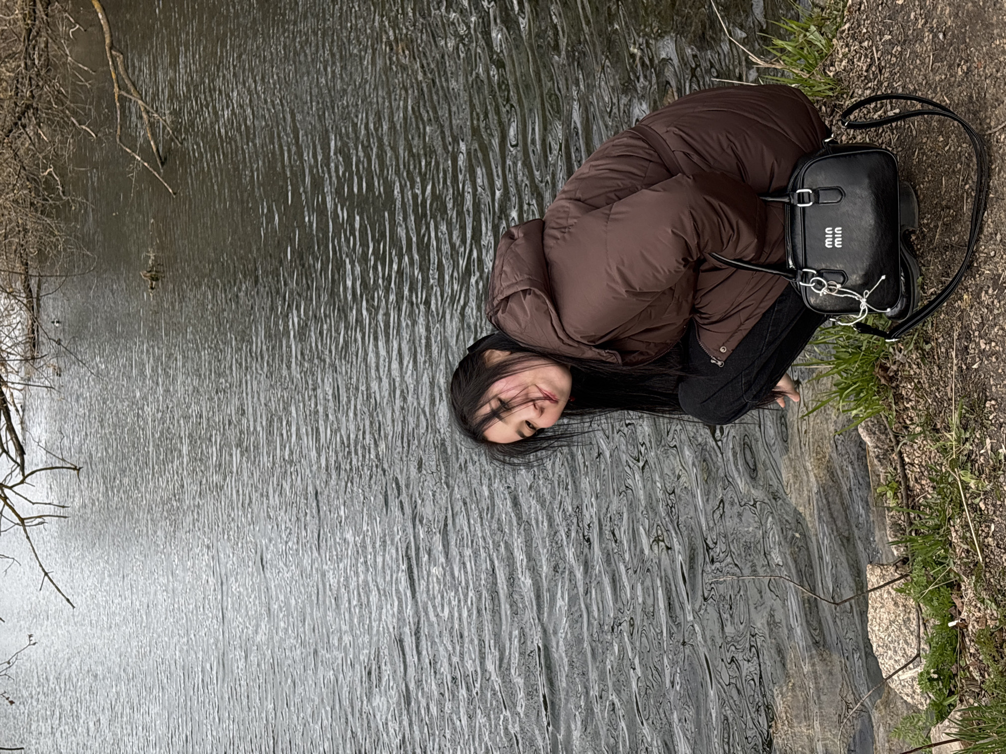 A young woman is depicted sitting or crouching by the edge of a body of water, likely a river or lake, in Kazan, Russia. She is positioned on the right side of the frame, on a bank composed of dirt, gravel, and sparse grass. Her long, dark hair falls across her face as she looks slightly upwards towards the camera. She is dressed in a brown puffer jacket and dark trousers, suggesting a cool climate.
Next to her, on the ground, is a black handbag with the white text "MIUMIU" visible on its front. The body of water occupies the majority of the image, featuring numerous ripples that create a textured, wavy surface, possibly due to a light breeze or current. In the distance on the water, a small duck is faintly visible. Bare tree branches extend into the frame from the far bank on the left, indicating either late autumn, winter, or early spring. The lighting appears soft and diffused, characteristic of an overcast day. The scene conveys a moment of quiet contemplation or relaxation amidst a natural, somewhat unkempt outdoor setting.