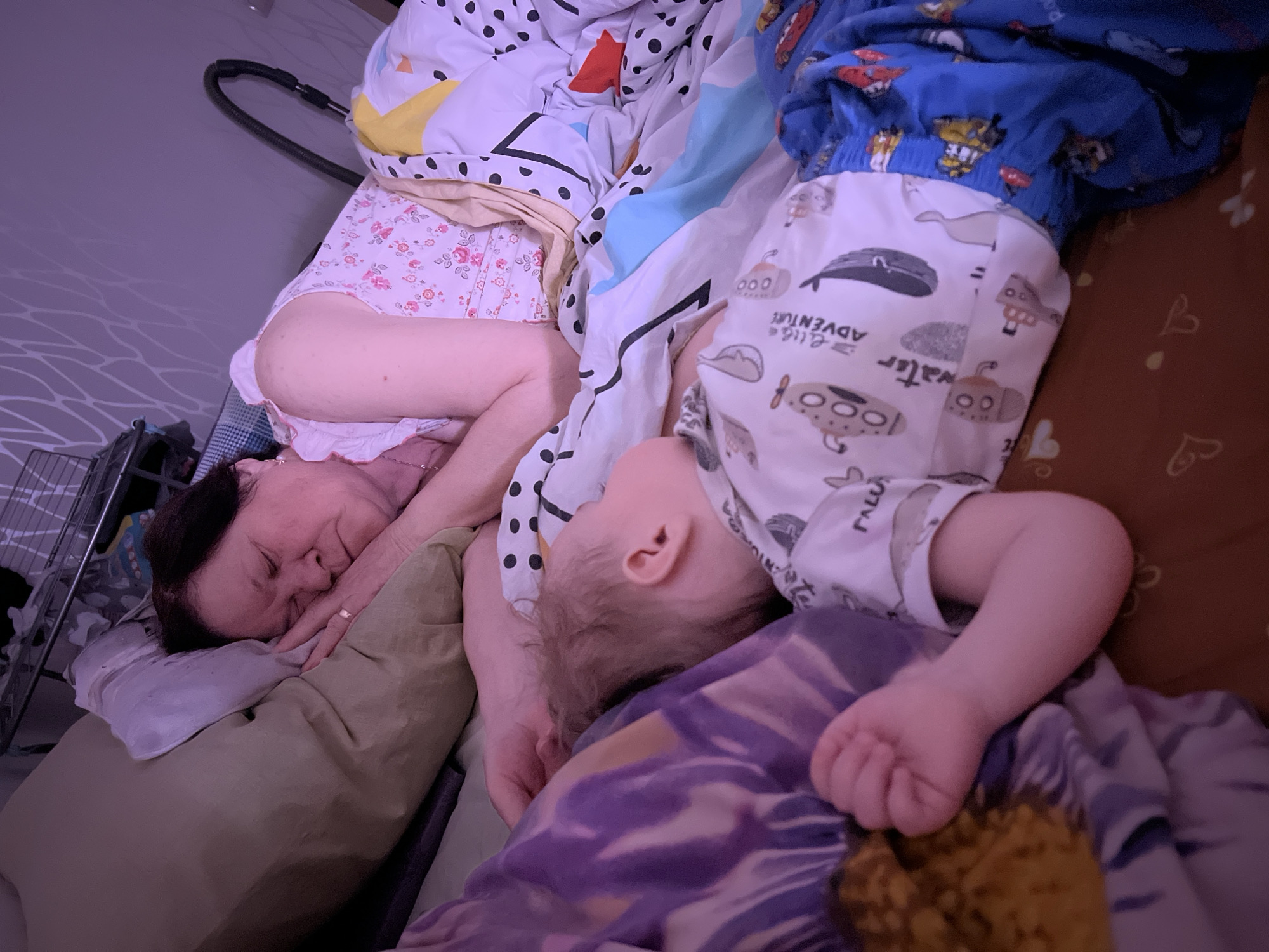 An intimate, dimly lit indoor scene captures an adult woman and a baby/toddler sleeping peacefully together in a bed in Perm, Russia. The image is taken from a slightly elevated, angled perspective.
The adult woman, with dark hair, is lying on her left side, her face gently pressed against a light green pillow. Her eyes are closed, indicating deep sleep. She wears light-colored sleepwear, part of which has a floral pattern. A ring is visible on her left ring finger. Her arm is positioned near the baby.
The baby/toddler lies perpendicular to the woman, with their head resting near her arm. The child's light brown hair is visible. They are clad in patterned sleepwear: a white bottom piece features various grey and black cartoon motifs including submarines, whales, and the partially visible text "Water" and "ADVENTURE." The upper body is covered by blue sleepwear adorned with colorful cartoon characters. The child's small hand is clenched, suggesting comfort in sleep.
The bed is covered with multiple layers of bedding. One duvet has a white background with black polka dots and geometric shapes in yellow, blue, and red. Another piece of bedding is brown with a subtle heart pattern. A purple and patterned blanket is also visible, partly covering the baby.
In the background, to the left of the woman, a black flexible hose, possibly from a vacuum cleaner, is partially visible, along with a metal storage rack. The overall lighting is soft with a warm, slightly purple hue, contributing to the tranquil atmosphere of slumber. Both individuals appear calm and at rest.