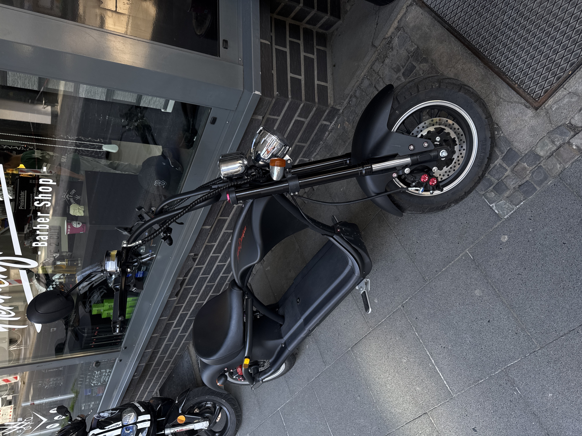 An image, rotated 90 degrees clockwise, captures an outdoor urban scene featuring a black electric scooter parked on a paved sidewalk in front of a barber shop. The scooter, a "fat tire" style, is the primary subject, displaying thick wheels, disc brakes with red calipers, chrome handlebars and headlights, and a black seat. A small orange indicator light is visible near the headlight. The name "DAZAISN" is visible on the tire sidewall.
In the background, the large glass windows and dark grey frame of the "Barber Shop" are prominent. White script text on the window clearly spells "Barber Shop." Other reflected text includes "Preisliste:" (Price list), "e CUT", "to go", "38", "RL", and partially visible names or words like "Haw" and "BL". Reflections in the shop window show elements from inside, including what appears to be a person, a baseball cap (possibly a Boston Red Sox cap given the "B" logo), and various barber supplies.
A second, smaller black scooter or motorcycle, partly obscured, is visible in the lower left corner, featuring white stripes and text that appears to read "HERO". The ground consists of grey rectangular paving stones and some smaller cobblestone-like sections, with a metal grate visible to the right of the main scooter's front wheel. The scene suggests a dry, daytime setting. No active event is taking place; both vehicles are stationary.