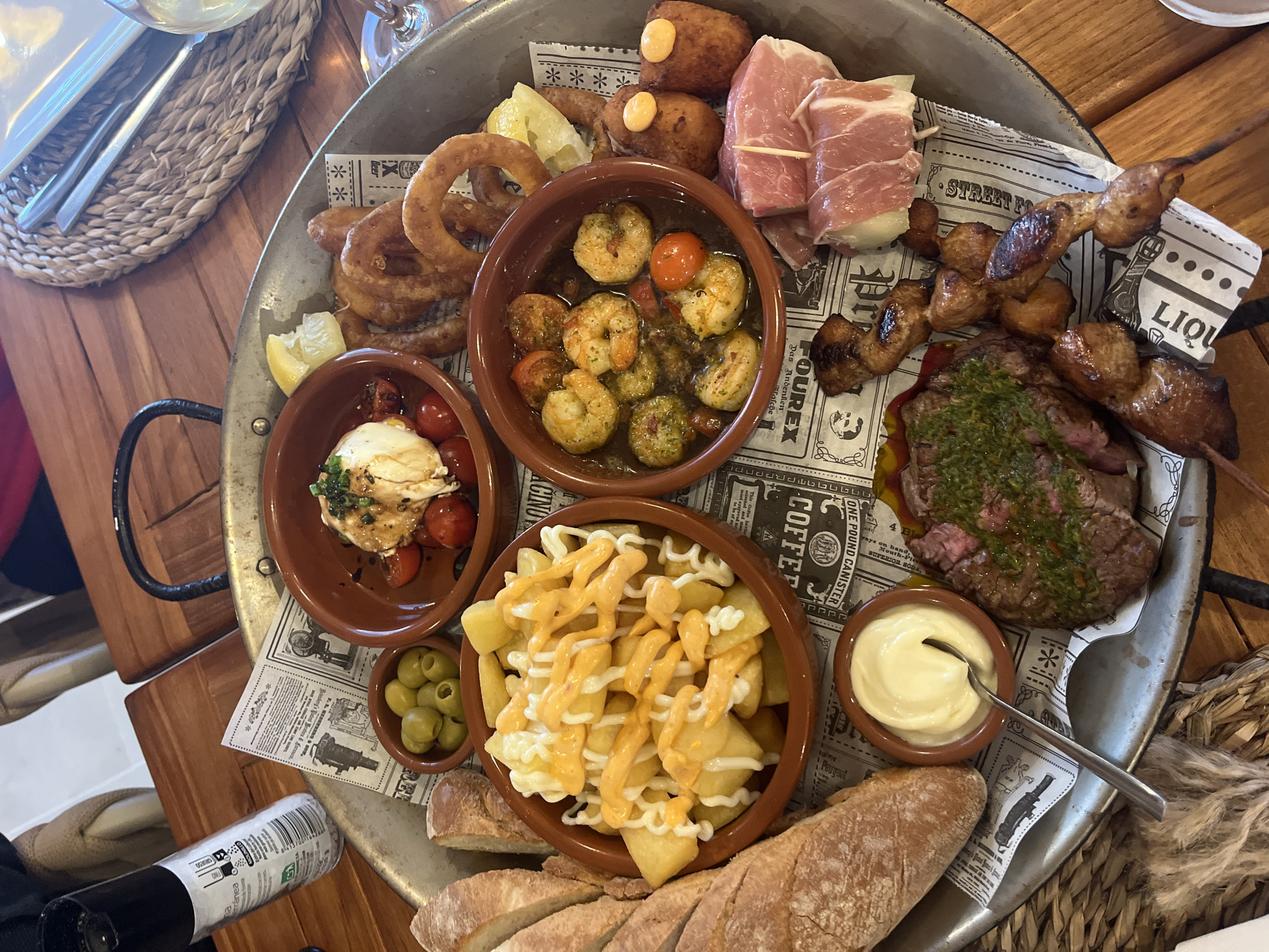 This image shows a variety of tapas arranged on a metal tray lined with newspaper print, likely served at a restaurant in Xàbia / Jávea, Spain. The tray contains various dishes, including fried calamari rings, shrimp with tomatoes in oil, croquettes, ham-wrapped melon, grilled meat skewers, a sliced steak with green sauce, patatas bravas, a small bowl of olives, bread, and a ramekin of mayonnaise. A silverware set rests on the edge of the tray, and a bottle and other dining accessories are partially visible on the wooden table around the tray.
