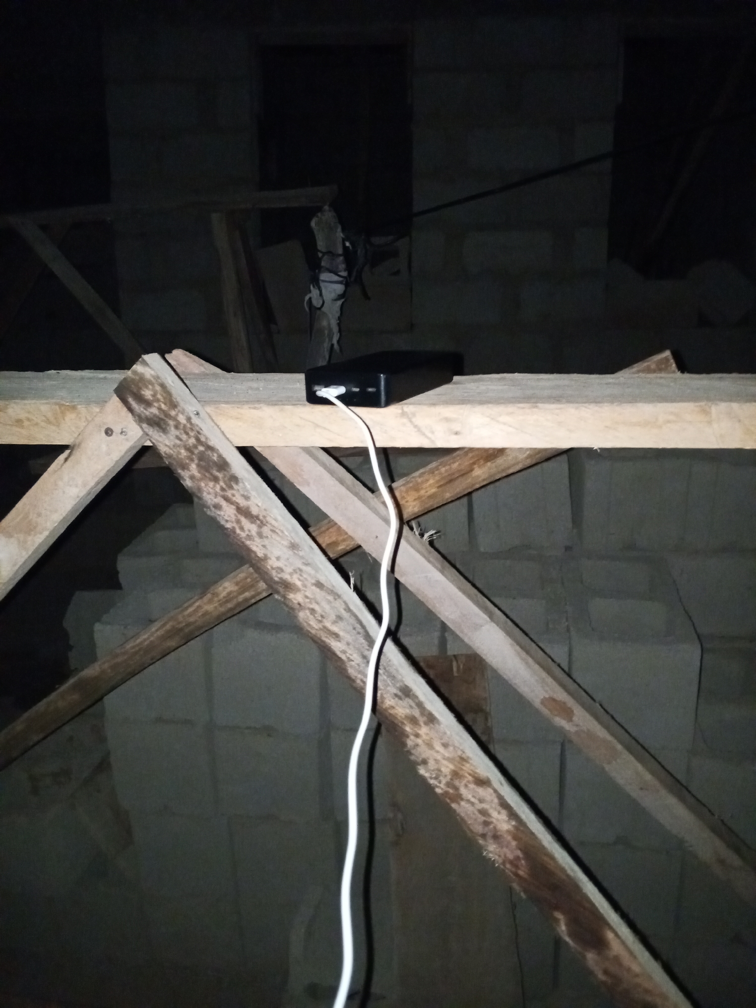 The image, taken at night, depicts a dark, unfinished construction site, likely in Bukuru, Nigeria. The foreground shows rough wooden scaffolding, appearing weathered and stained. On top of the scaffolding sits a black rectangular object with a white cable plugged into it. In the background are cinder block walls, partially constructed. The environment is dark, suggesting limited or no artificial lighting. There is an unidentified object hanging in the background.