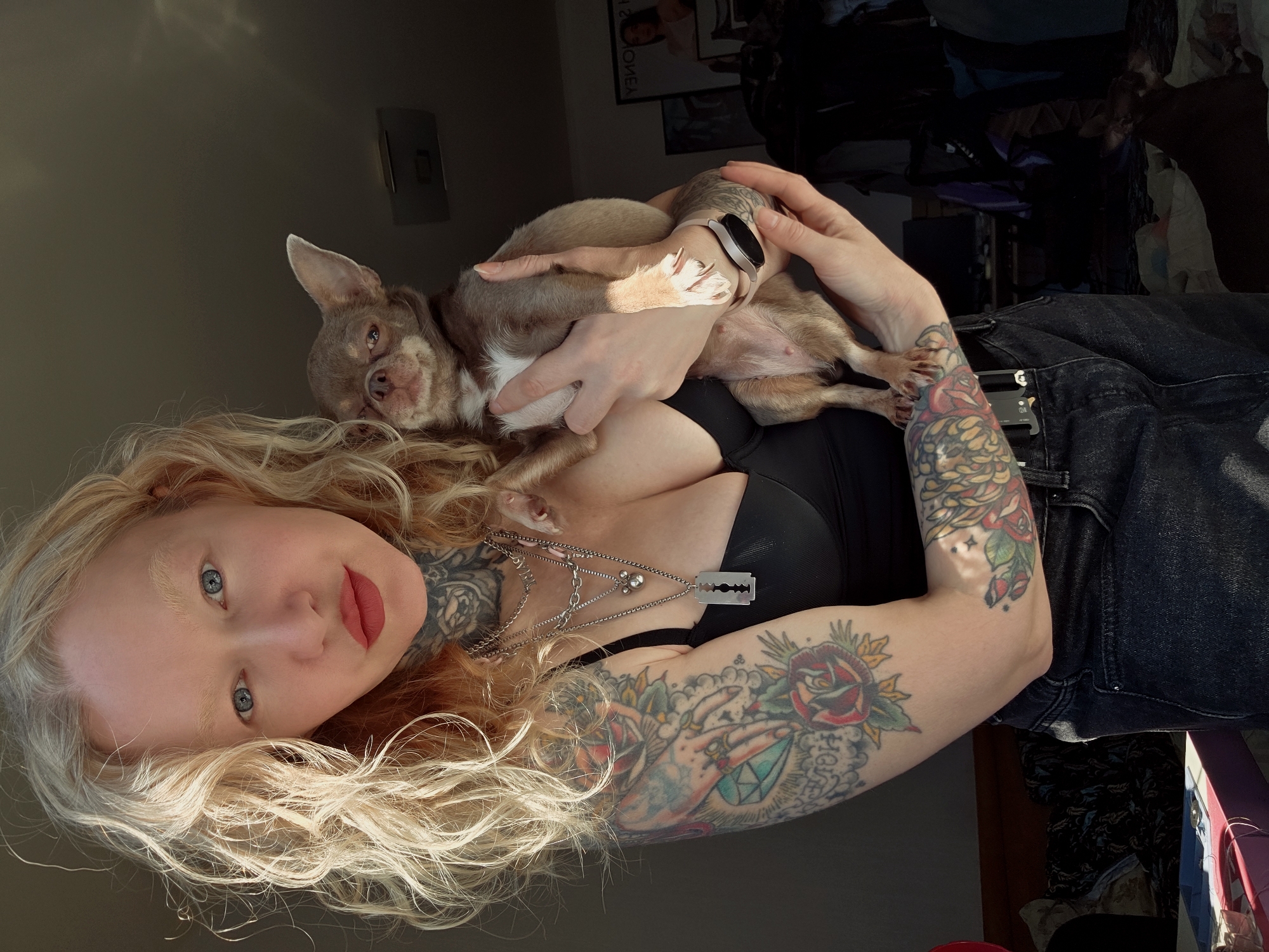A woman is holding a chihuahua. The woman has blonde hair, blue eyes, red lipstick, and tattoos on her neck and arms. She is wearing a black top and dark jeans. She is wearing a razor blade necklace. The dog is small and brown. The background is a room with a window and some furniture. It is impossible to determine if this is in Saint Petersburg, Russia.