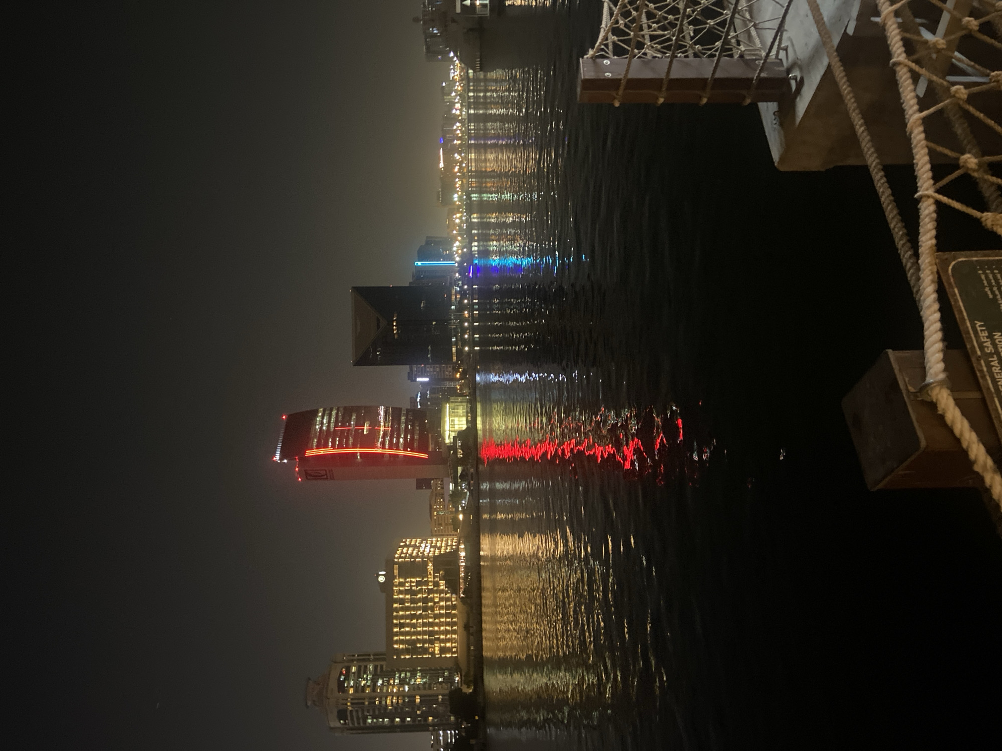 The image shows a nighttime view of the Deira Creek in Dubai, United Arab Emirates. The scene is dominated by the city skyline, illuminated by numerous lights reflecting on the dark water. In the foreground, a portion of a pier is visible with wooden posts and rope railings. Several buildings, including skyscrapers with distinct architectural designs and lighting, are silhouetted against the night sky. Notably, a tall building with red vertical lighting stands out. The water is calm, with ripples breaking up the reflections of the city lights. The overall atmosphere is urban and tranquil.