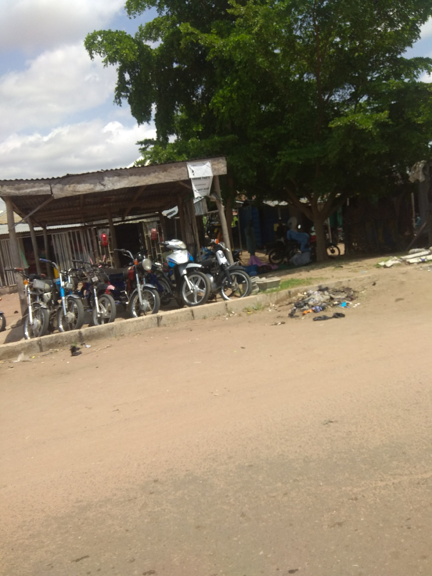 The image depicts a street scene in Jalingo, Nigeria. In the foreground, a dirt road is visible, with a curb running along its edge. A pile of trash, including plastic bags and what appear to be shoes, is scattered on the road. A line of motorcycles is parked along the curb, under a wooden shelter with a corrugated metal roof. The motorcycles are varied in color and style, with some appearing to be for sale or rent. Behind the motorcycles, a dense green tree provides shade. In the background, there are various stalls and structures, with some people visible, though their actions are unclear. The sky is partly cloudy, with white clouds against a blue background. The overall impression is of a busy, informal marketplace or transport hub.