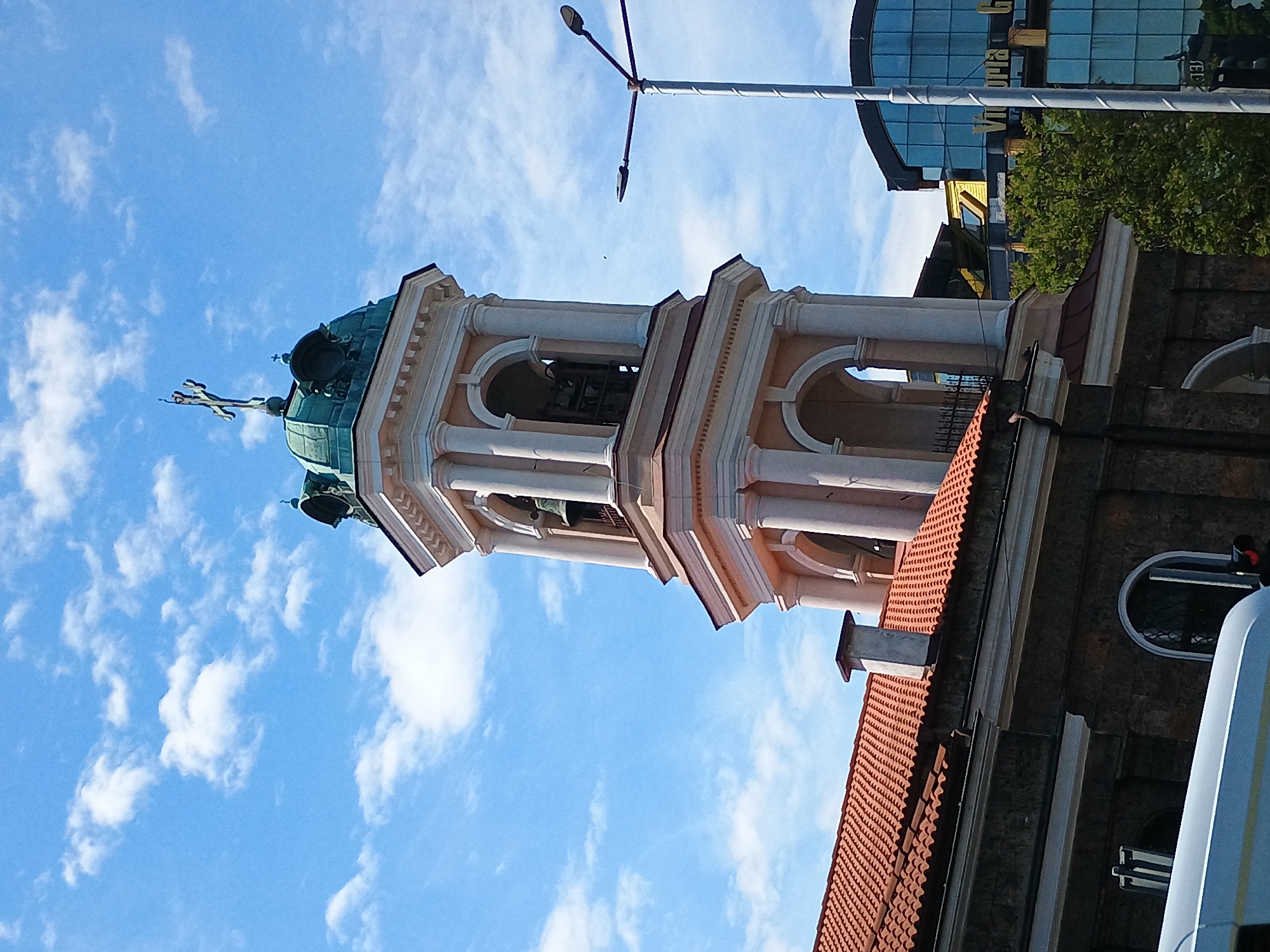 The image depicts a church tower in Unknown, Unknown, featuring a green-domed cupola topped with a cross. The tower has an open structure supported by white columns, with arches revealing bells inside. The tower's architecture exhibits ornate details. Below the tower, a red-tiled roof is visible. A street light is positioned near the tower, and a modern glass building rises in the background. The sky is blue with scattered white clouds.