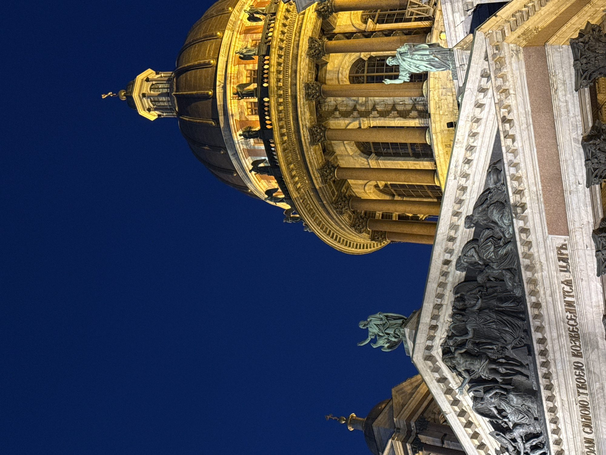 This image presents a dramatic, upward-angled night view of a grand cathedral, strongly resembling Saint Isaac's Cathedral in Saint Petersburg, Russia. The immense structure is illuminated by warm, golden artificial lights, contrasting sharply with the deep, uniform blue of the night sky.
The most prominent feature is the cathedral's majestic, gilded dome, which gleams brightly at the top of the frame. Below it, a large cylindrical drum supports the dome, featuring classical columns and arched windows. Numerous statues of figures, appearing in a teal or verdigris hue, are positioned along the drum and on various ledges of the building, adding to its ornate decoration.
The building's white stone facade is richly adorned with intricate sculptural reliefs, depicting multiple figures in dynamic compositions. Below these reliefs, a line of Cyrillic text is clearly visible, inscribed on the stone. The text reads: "ОДАЙ СИЛОЮ ТВОЕЮ ВОЗВЕСЕЛИТСЯ ЦАРЬ." This translates to "The king shall rejoice in Your strength," a biblical verse commonly associated with Psalm 21.
The low vantage point emphasizes the cathedral's towering height and architectural grandeur. The artificial lighting beautifully highlights the intricate details, rich textures, and the overall monumental scale of the building, creating an awe-inspiring and majestic scene during twilight or nighttime in Saint Petersburg.