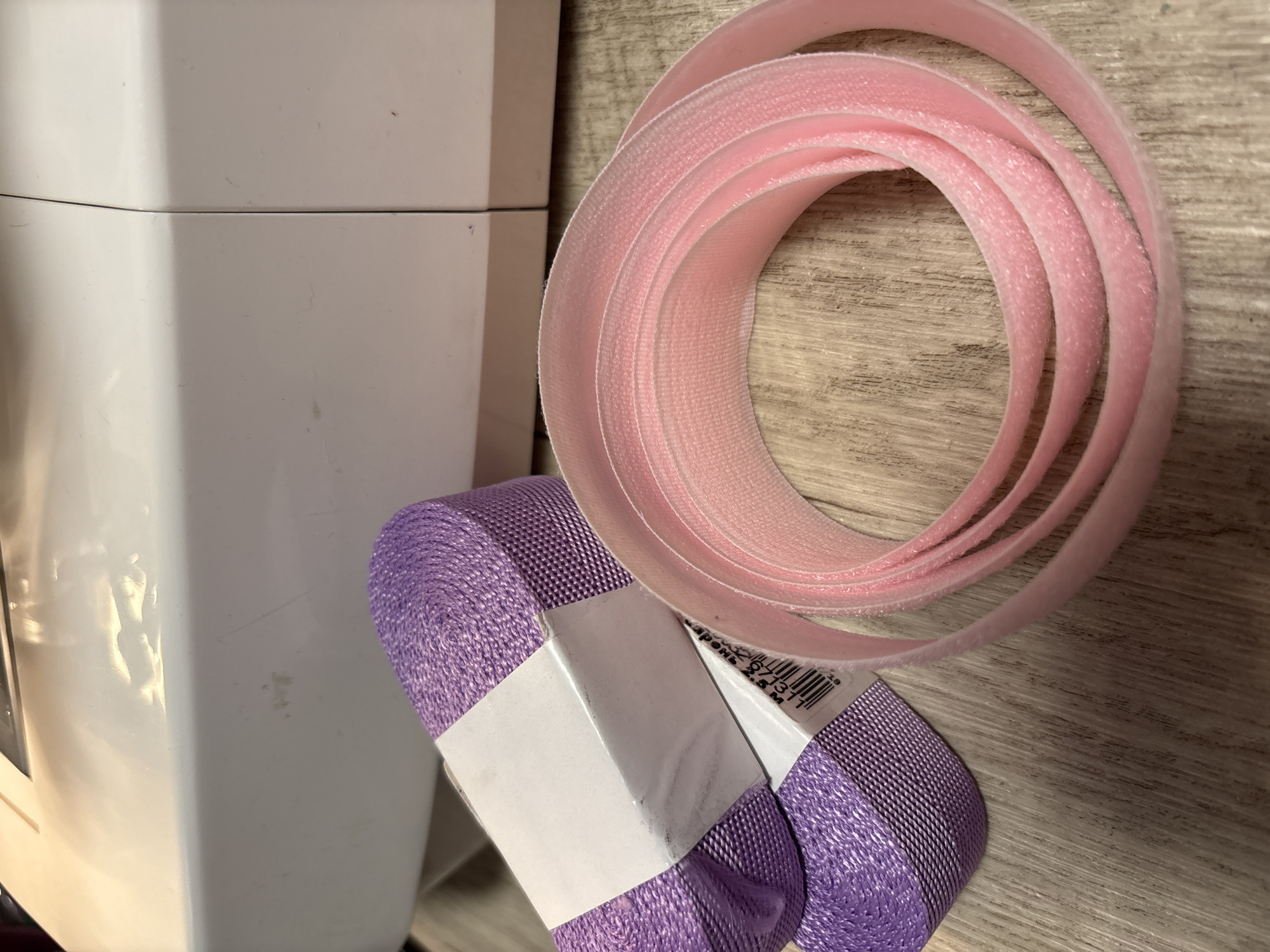 The media file presents a close-up, overhead view of crafting supplies laid out on a light-colored wooden surface with a visible grain. The scene is indoors, likely in a domestic or workshop setting.
In the foreground, two rolls of purple webbing or heavy-duty ribbon are stacked horizontally. Each roll is bound by a white paper label, one of which displays a barcode and partially visible text. The discernible text on the label includes "2.5 M," indicating a length of 2.5 meters. Other fragments of text, likely in Cyrillic, are visible, such as numbers like "971311" and a word that could be "ЛЕНТА" (ribbon/tape).
Above these purple rolls, a large coil of pink hook-and-loop fastener material, commonly known as Velcro, is prominently displayed. The pink material has a soft, textured surface characteristic of the loop side of the fastener. To the left of the frame, a portion of a white appliance or container is visible, appearing sleek and unadorned.
No people are present, and no direct activity is taking place, but the arrangement of materials suggests a preparation for, or the aftermath of, a crafting, sewing, or DIY project. The lighting indicates an indoor environment, with no specific time of day or weather conditions discernible. The overall impression is one of organized supplies in a functional space, potentially in Прикамский, Russia, given the provided context.