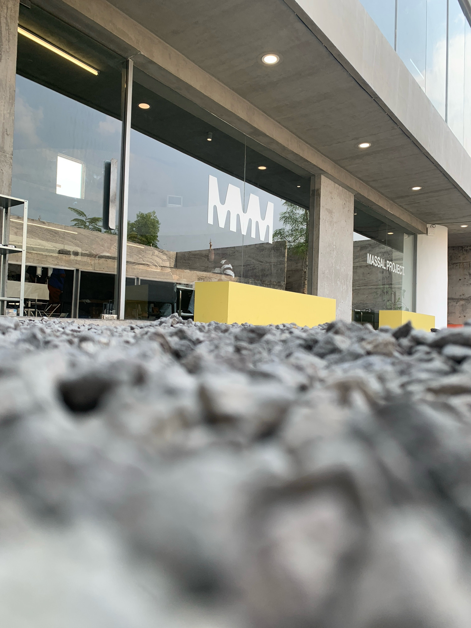 The image shows the exterior of a building with a modern architectural style, characterized by concrete elements, large glass panels, and minimalist design. In the foreground, a layer of rough grey gravel is visible, with the camera positioned very low to the ground, creating a perspective that emphasizes the texture and scale of the gravel.
Through the glass facade, the interior of the building is partially visible. There are reflections on the glass, showing trees and the sky. A large white logo, possibly an "MM," is displayed on the glass. Further along the building, the text "MASSAL PROJECT" is also visible on the glass.
Inside, a bright yellow rectangular structure is positioned against the glass, possibly a bench or display element. In the background, some individuals can be seen inside the building, one wearing an orange-brown garment. The overall environment suggests a public space, potentially a gallery, showroom, or a commercial establishment. The context provided places this scene in the city of Meri, Indonesia.