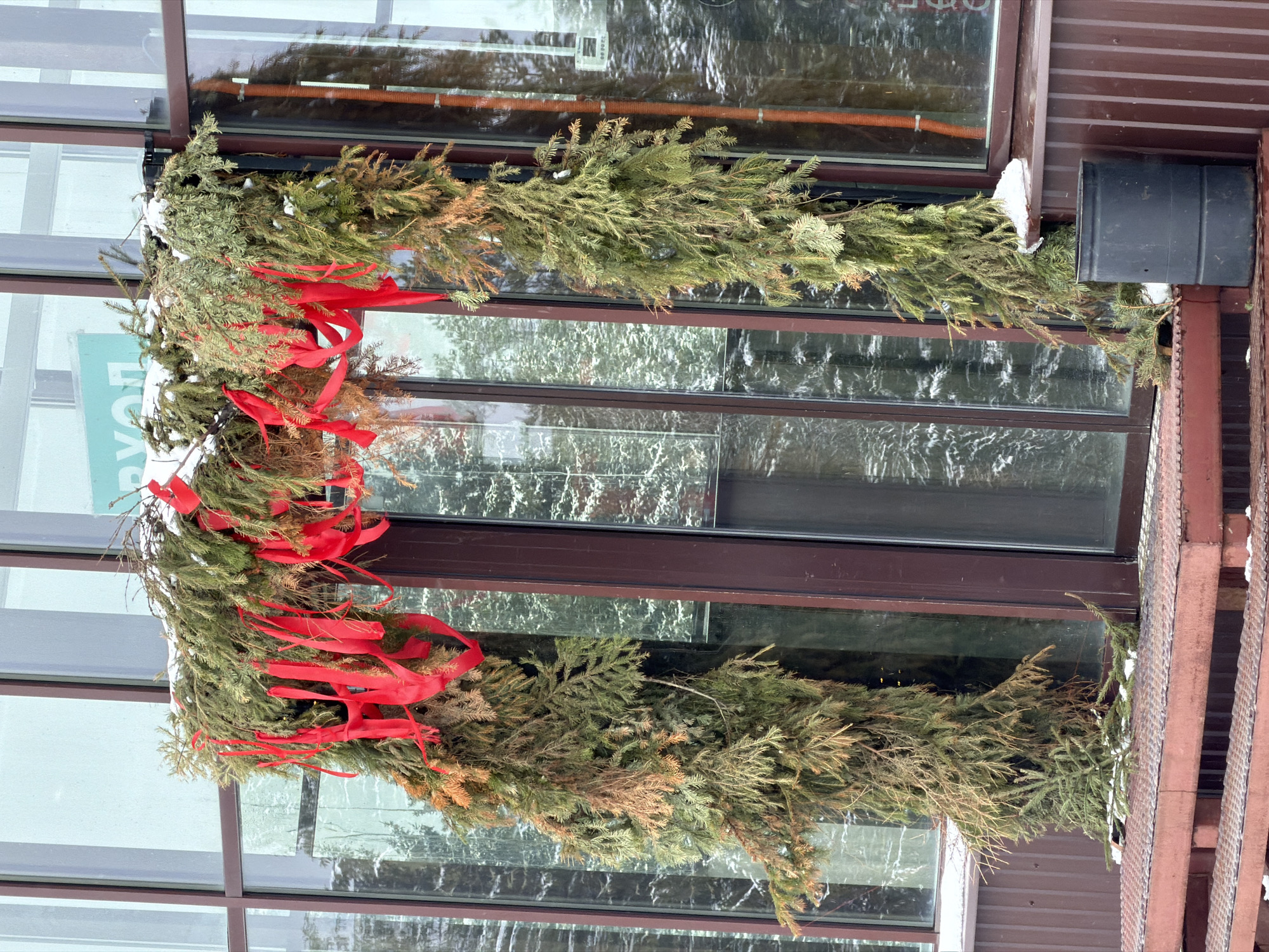 The image depicts a Christmas decoration on a building in what is likely Нечкино, Russia. The decoration consists of evergreen branches, possibly fir or spruce, adorned with red ribbons, arranged in a semi-circular garland or swag. The garland is mounted on a window frame with multiple panes. The building has a brown-colored exterior trim, and there is a metal staircase or framework visible to the right. The reflection in the window panes suggests an outdoor setting, possibly with trees and a cloudy sky.