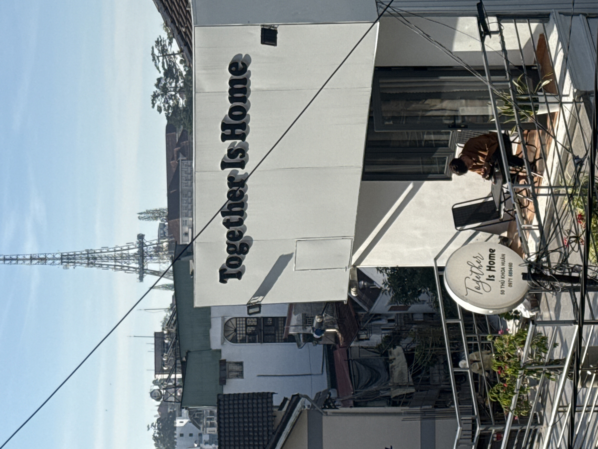 The image shows a building with the text "Together Is Home" prominently displayed on its exterior. The building appears to be in an urban or semi-urban setting, with other buildings and structures visible in the background, including a tall tower that resembles a radio or television antenna. In the foreground, a person is seated at a table on a balcony, looking down at their hands. There are chairs and potted plants on the balcony. A circular sign with the text "Together Is Home" and an address "50 THU KHOA HUAN" and phone number "0971 689460" is also visible. The overall scene suggests a commercial establishment, possibly a cafe or accommodation, in Da Lat, Vietnam.