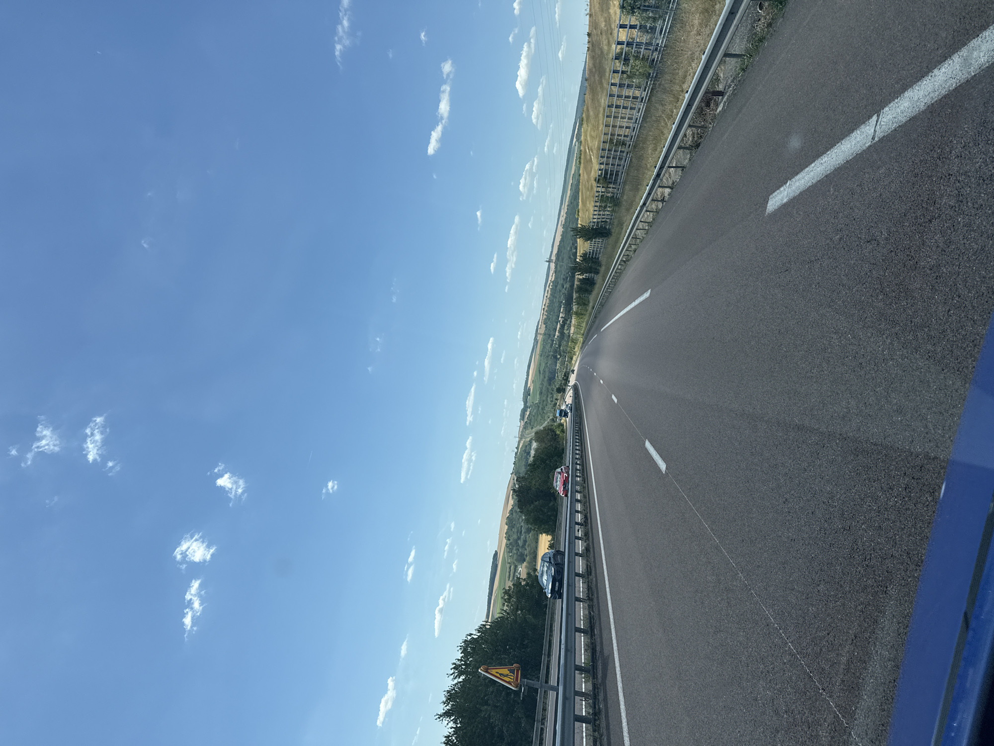 The image shows a view from a vehicle traveling on a highway in the rural area surrounding Bouleuse, France. The sky is bright blue with scattered white clouds. The highway has asphalt pavement and is divided by white lane markings. Several cars are visible on the road. On the left side of the highway, there is a guardrail, trees, and fields. On the right side, there is a metal fence, dry grass, and trees in the distance. A yellow warning sign with a depiction of a person crossing the road is visible on a pole on the left side of the highway. The landscape is hilly and appears to be agricultural or natural terrain.