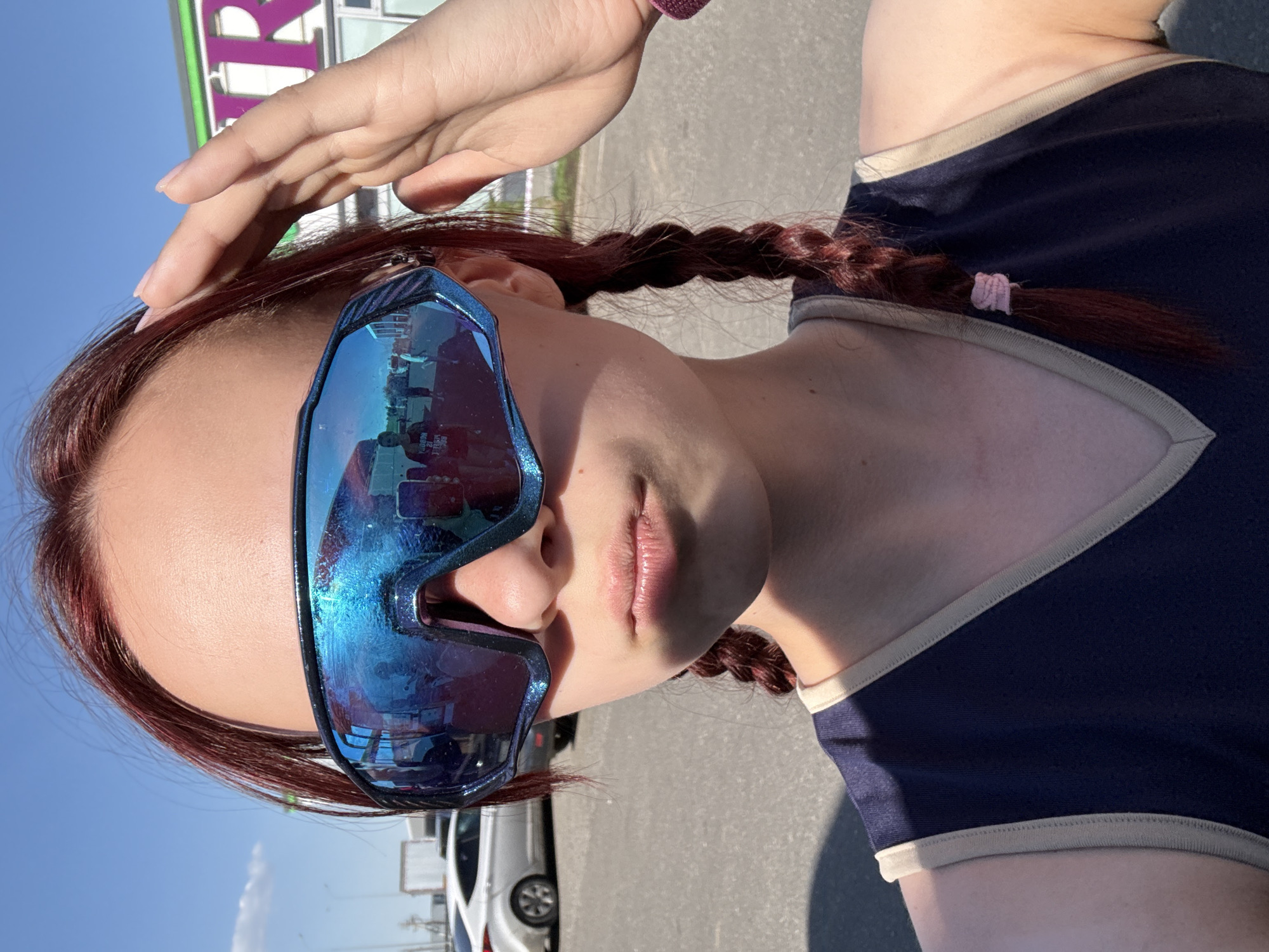 A woman is standing outside on a sunny day, with a light-colored sky visible in the background. She has auburn hair styled in braids. She is wearing large, reflective blue sunglasses that obscure her eyes. Her left hand is raised towards her forehead. She is wearing a dark blue top with light-colored trim. Cars are parked in the background. The location is Казань, Russia, as identified by a sign with Cyrillic characters. The setting appears to be a parking lot or street near a commercial building.