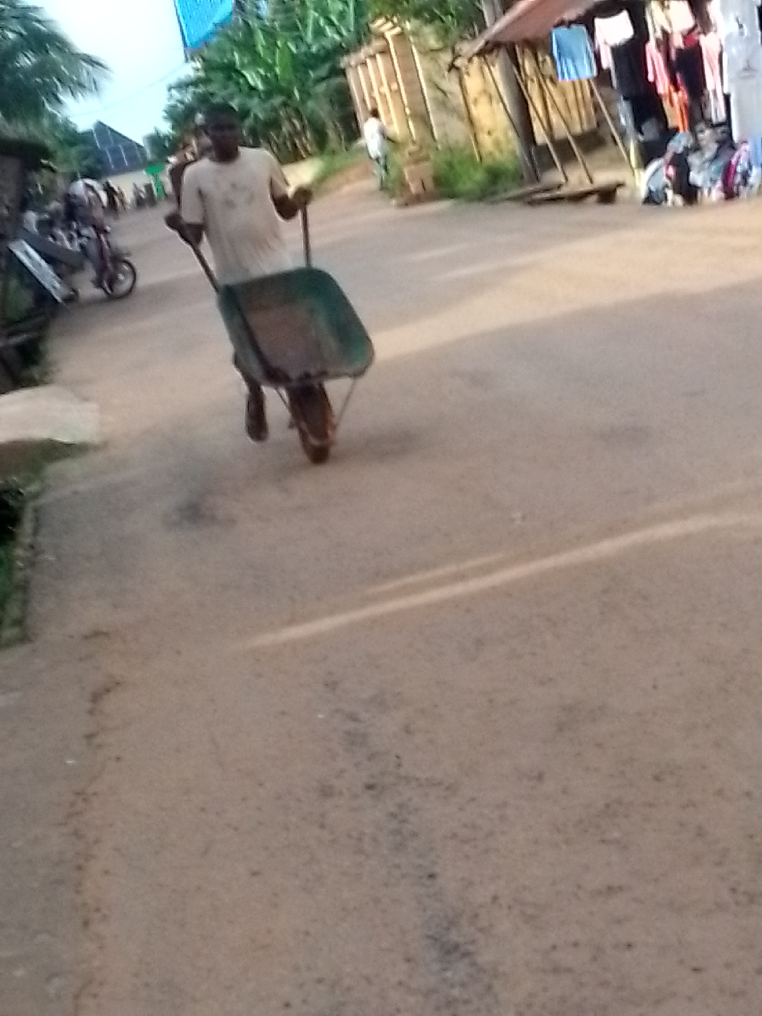 A man is pushing a wheelbarrow along a dirt road in Orlu, Nigeria. He is wearing a light-colored t-shirt. To the right of the road is a rudimentary stall selling clothes. A motorcycle is parked in the distance to the left. Vegetation surrounds the scene.