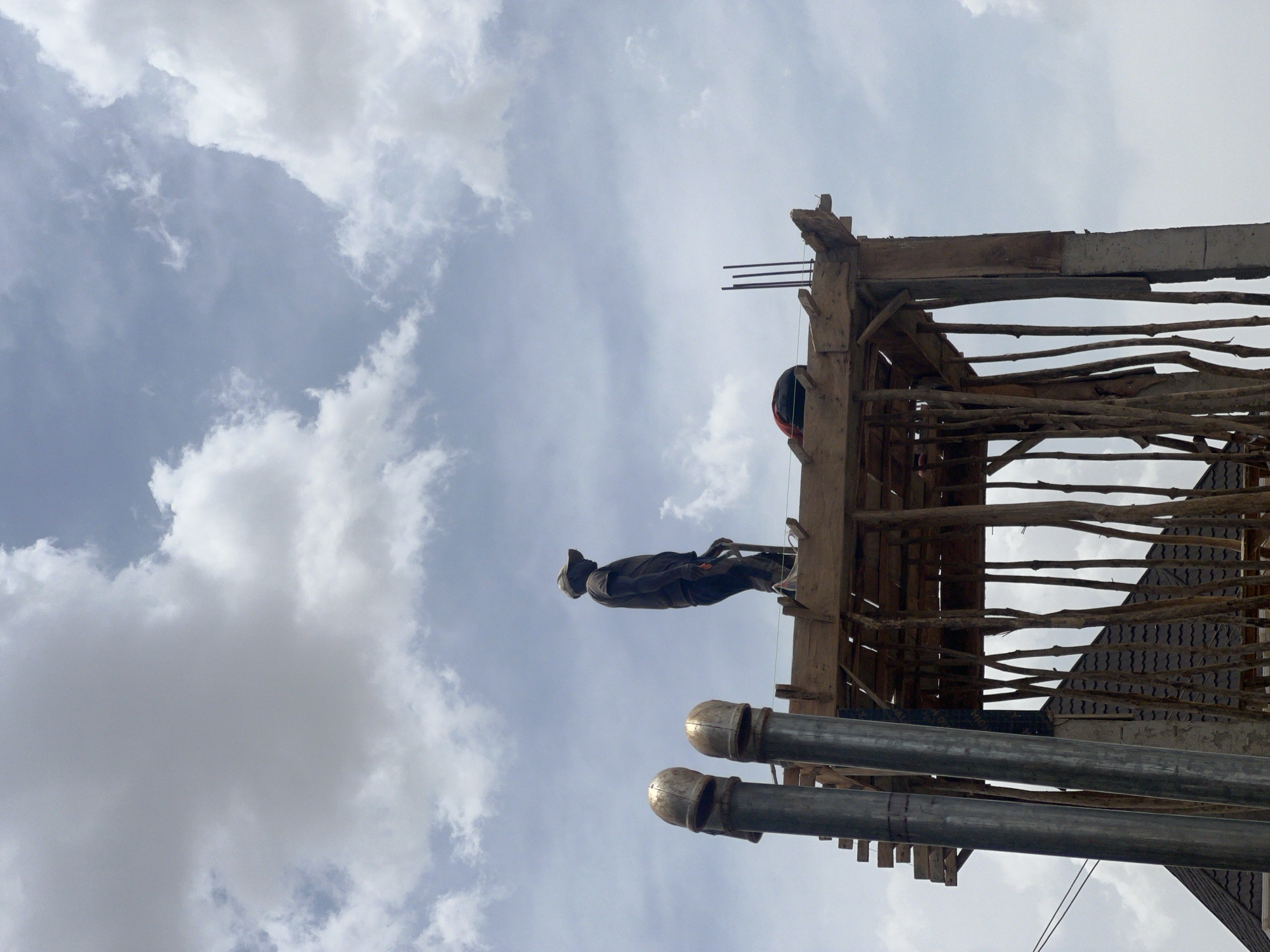 From a low angle, the image shows a construction worker on a wooden structure with the sky as the backdrop in Jos, Nigeria. The worker wears dark clothing. Below the structure are pipes. The sky is overcast with patches of blue.