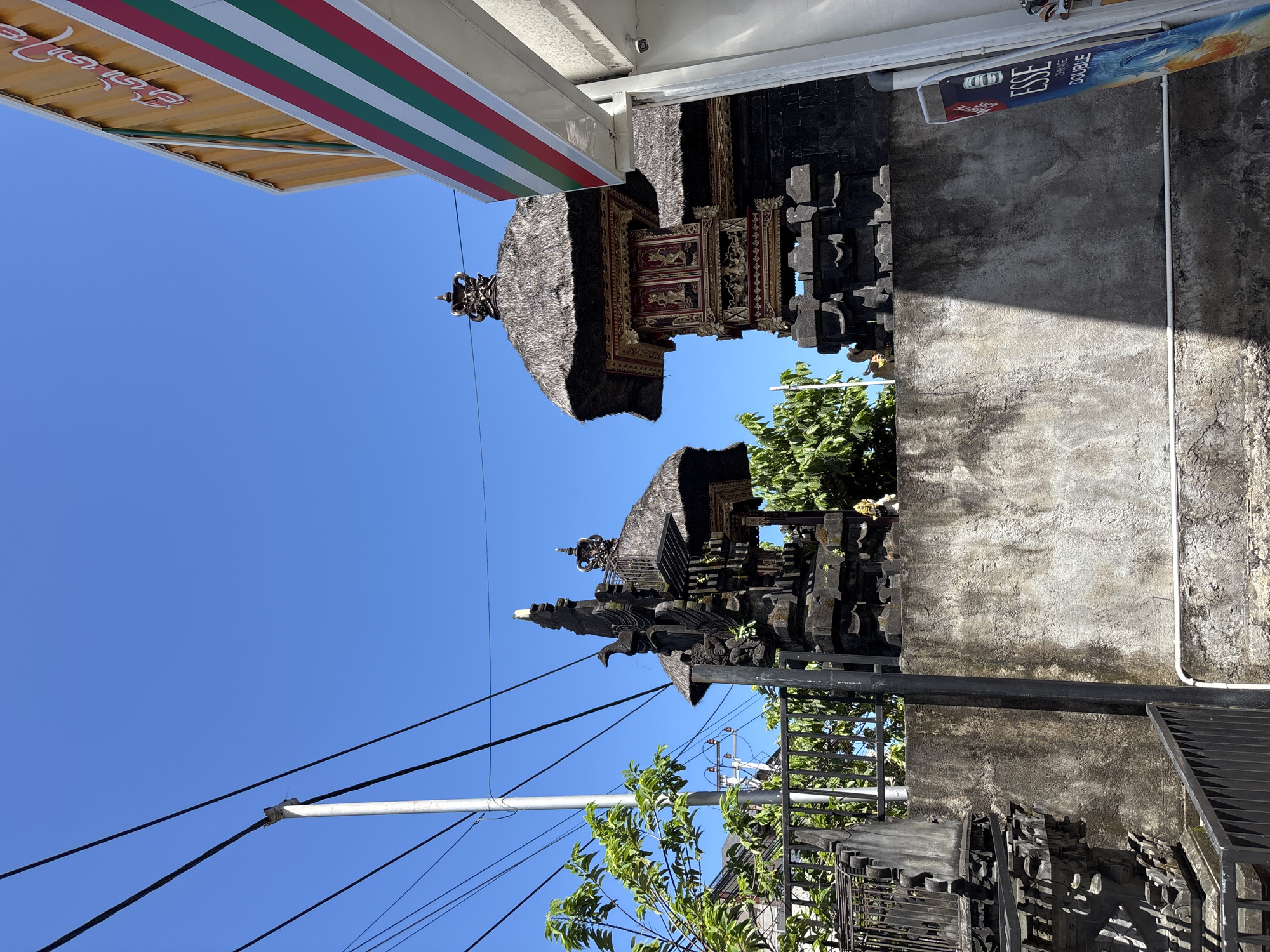 Captured in Kuta Selatan, Indonesia, the image presents a juxtaposition of traditional Balinese architecture and modern commercial elements. Two Balinese shrines or temples are prominently featured, displaying tiered structures with thatched roofs and intricate carvings. Adjacent to these traditional structures is a modern building, identifiable by a commercial awning with red, white, and green stripes and the visible signage of a convenience store. A wall made of grey, weathered material sits next to a sign advertising ESSE cigarettes. Utility poles and power lines cut across the blue sky, adding to the mix of cultural and modern elements. The scene appears to be a street view, capturing the blend of local culture with commercial development.