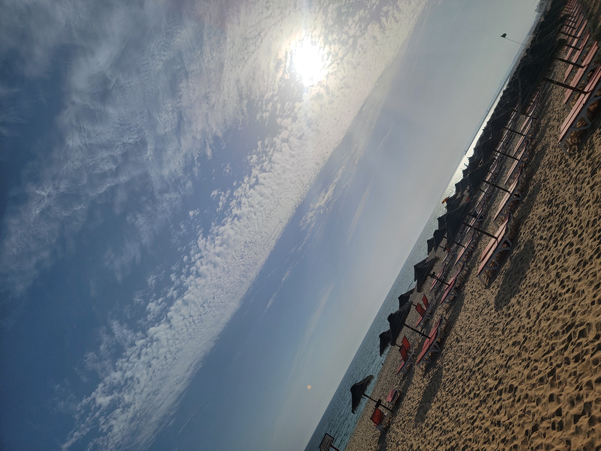 This is a photograph of a beach scene in Quarteira, Portugal. The image is taken from a low angle, looking diagonally across the sandy beach towards the sea. In the foreground, the sand is textured with ripples and the shadows of beach umbrellas and lounge chairs. A row of red lounge chairs with dark umbrellas are arranged in neat rows, suggesting a prepared beach for visitors. The sea is visible in the middle ground, appearing calm with a slight shimmer from the sun. The sky dominates the upper portion of the image and is filled with a dramatic display of clouds. The sun is bright and high in the sky, partially obscured by wispy, white clouds that stretch across the frame, creating a textured, almost painterly effect. The overall impression is of a bright, sunny day at the beach, likely in the late morning or early afternoon given the sun's position and intensity. There are no people visible in the frame. The image evokes a sense of tranquility and relaxation, typical of a seaside vacation. The location cues are consistent with a beach resort area.