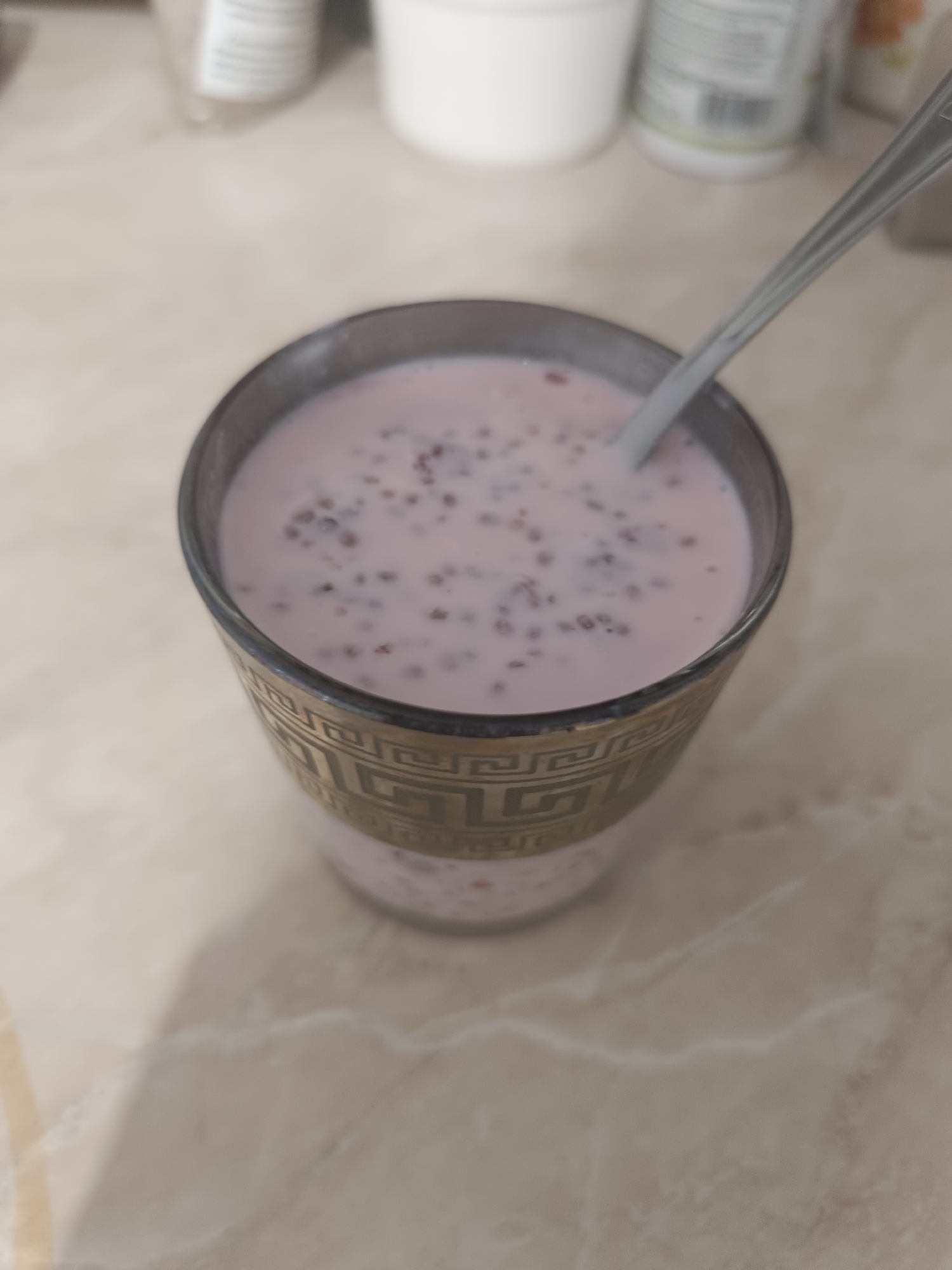 A close-up, high-angle shot shows a glass of pink liquid with small, dark specks, possibly seeds or berries, and a spoon submerged in it. The glass has a decorative gold band around its base. The surface the glass rests on appears to be a marbled countertop. In the background, out of focus, are some white containers and blurry objects, suggesting a kitchen or storage area. The lighting is diffuse, with no strong shadows. The specific location in the city of Taraz city administration, Kazakhstan, is not discernible from the visual information provided in the image.
