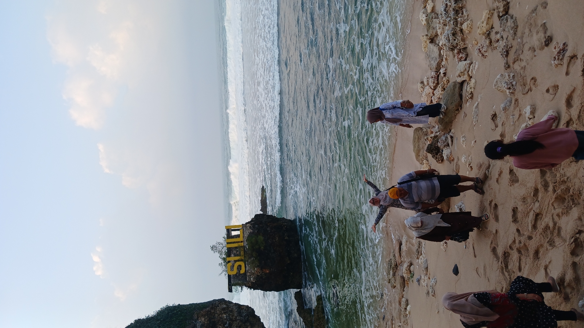 In the city of Tepus, Indonesia, a group of people are gathered on a sandy beach with scattered rocks near the water's edge. The ocean displays moderate waves breaking onto the shore under a partly cloudy sky. To the left, a rocky outcrop is visible, topped with a yellow sign that reads "SIULI." One individual in the foreground is seated on the sand, facing away from the camera. Further back, several other individuals are standing and walking on the beach. One person in the center is posing with arms outstretched, while another stands to their right. Two individuals are walking together further back, and another is standing on a rock formation to the right of the frame. The overall scene captures a casual outing at a coastal location.