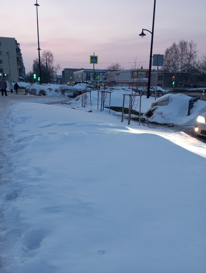 The image presents a snow-covered urban landscape in Yaromaska, Russia, observed during twilight hours. The foreground is dominated by undisturbed snow, forming what appears to be a pedestrian path, with isolated footprints visible. To the immediate left, a cleared section of the path shows four individuals, dressed in dark winter clothing, walking away from the camera. In the background on the left, a multi-story residential building with a light-colored facade is visible. A tall street lamp stands near this building, and a traffic light displays a green signal.
Towards the center and right, a series of young, bare trees, each supported by wooden stakes, line a snow-covered median. Large, irregular mounds of snow, likely accumulated from plowing, are present. A black, ornate street lamp extends over the street on the right, illuminating a portion of the snow. A white vehicle, with its headlights illuminated, is partially visible in the bottom right corner, indicating its presence on the roadway. Directly across the street, a yellow pedestrian crossing sign is prominent. Behind it, a commercial building features a red and white facade with the Cyrillic text "МЕБЕЛЬ" (Furniture) clearly visible. Further down the street on the right, another traffic light displays a red signal. Deciduous trees, devoid of leaves, are scattered throughout the background. The sky exhibits a pale pink and light blue gradient, characteristic of dawn or dusk.