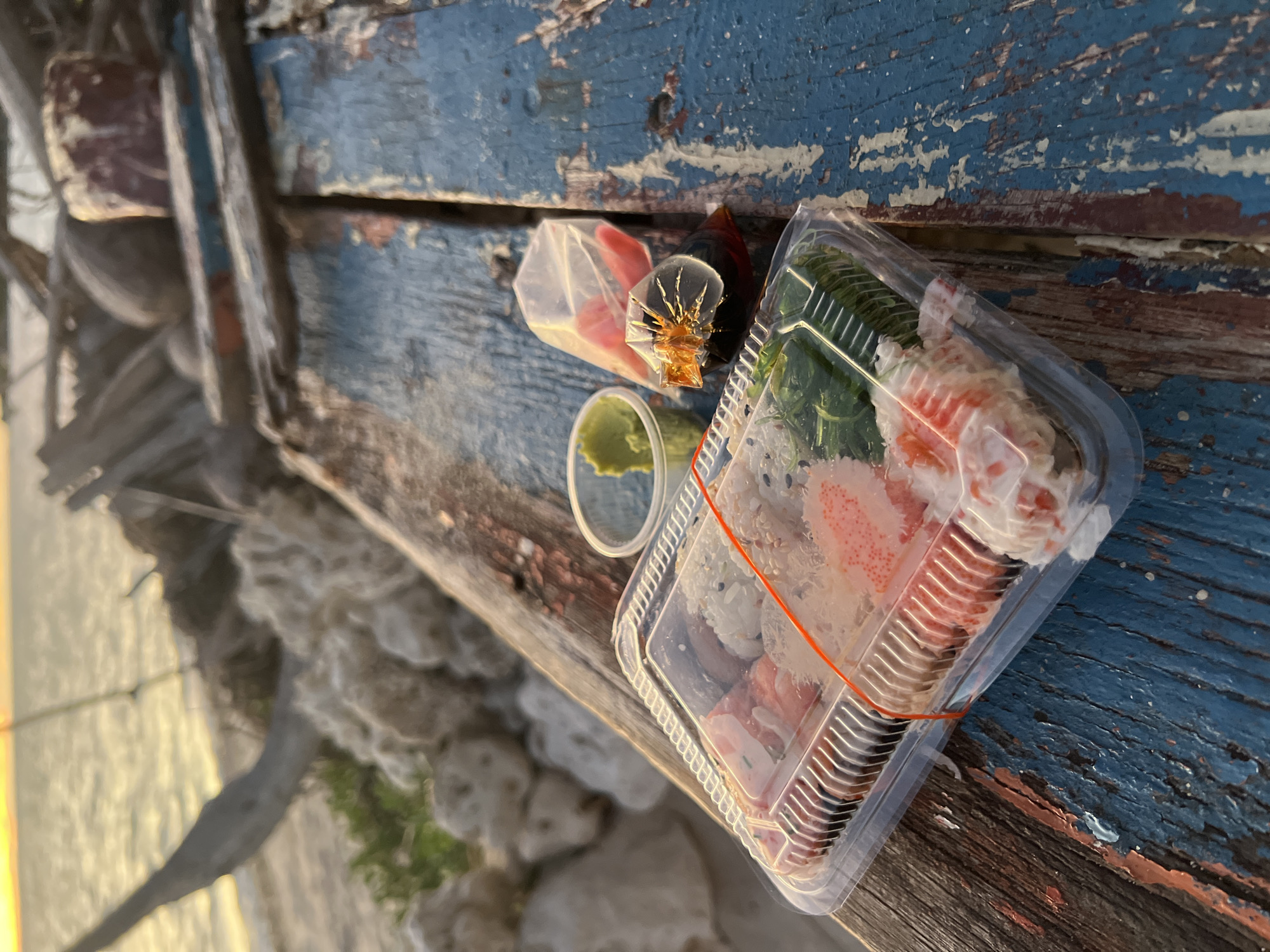 A clear plastic container of sushi sits on a weathered, blue-painted wooden surface. The sushi includes rolls with orange roe, white rice, and green seaweed. Next to the sushi, in small plastic bags, are packets of soy sauce and pickled ginger, and a small container of green wasabi. In the background, the ocean and beach are visible, with driftwood and rocks on the sand. The scene appears to be on a beach in Koh Phangan, Thailand, possibly during sunset due to the warm lighting.