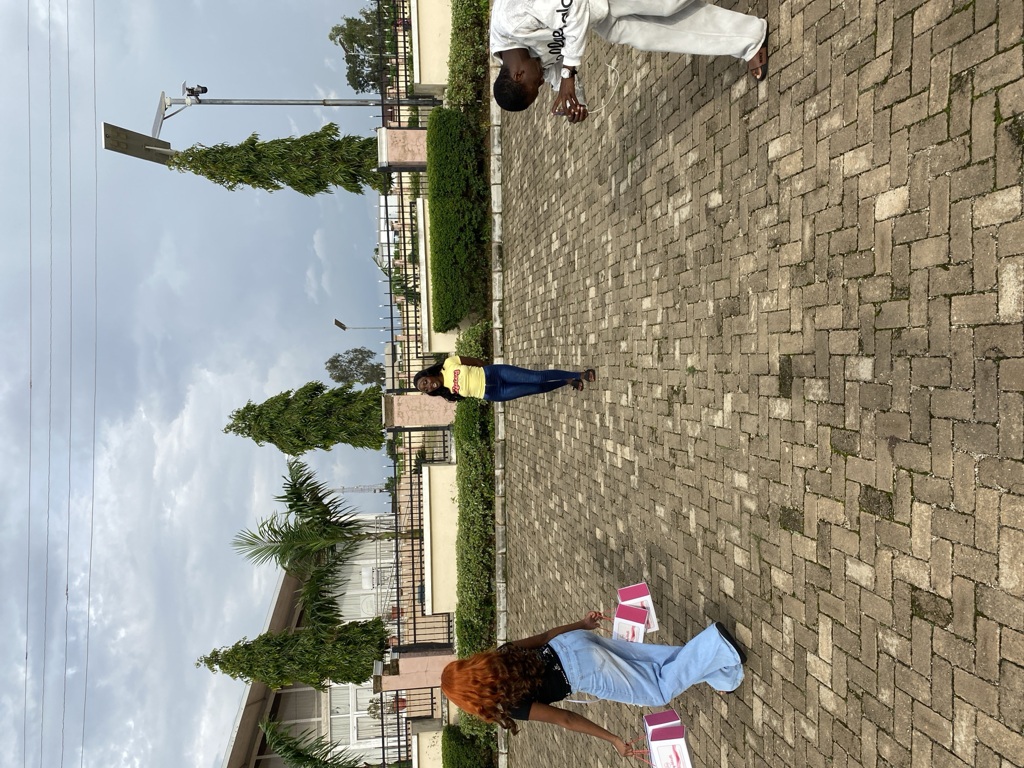 In an outdoor paved area, three individuals are present. In the foreground, a person with bright orange hair, wearing a black t-shirt and light blue wide-leg jeans, walks away from the camera holding two pink and white gift bags. To their left and further back, a woman in a yellow t-shirt with the word "spices" printed on it and dark blue jeans stands facing the camera, smiling. To the right, a person in a light gray tracksuit and slides is bending down, appearing to interact with a wire on the ground. In the background, a modern building with large windows and a fence is visible, with several tall green trees and palm trees bordering it. The sky is overcast with clouds. The scene is located in Barakin Lerawi, Nigeria.