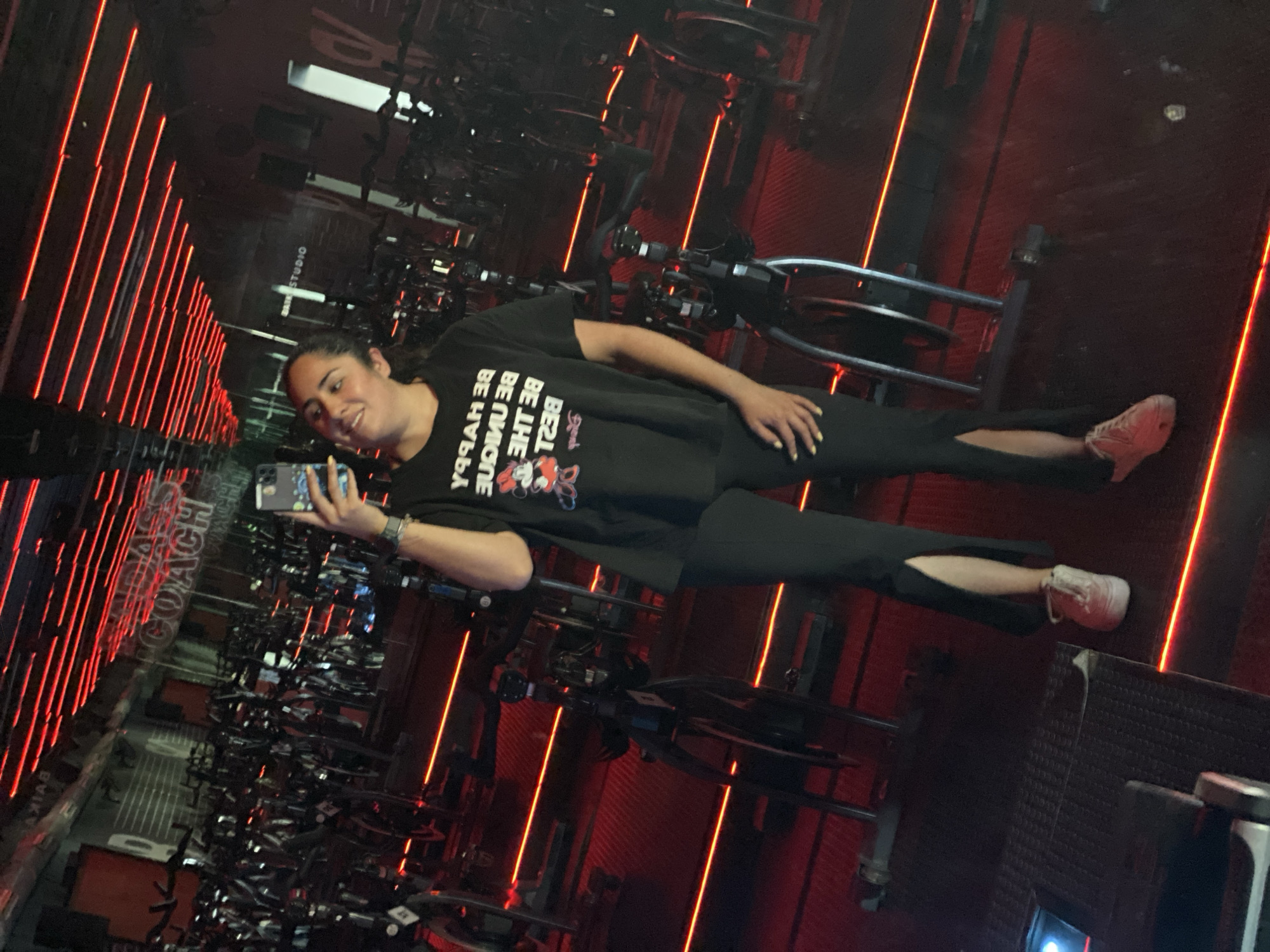 A woman is standing in a dark room with rows of exercise bikes. The room is lit by red neon lights. The woman is wearing a black t-shirt with an illustration of Minnie Mouse and the words "Be Happy Be Unique Be Yourself". She is also wearing black pants and pink sneakers. She is holding a cell phone and appears to be taking a picture of herself. It is not possible to determine the specific location in Puebla, Mexico from the given image.