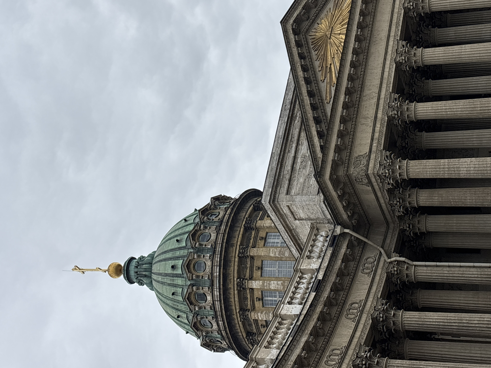 The image shows a low-angle view of the Kazan Cathedral in Saint Petersburg, Russia, against an overcast sky. The cathedral's dome, adorned with a golden spire and cross, is visible on the left side of the frame. To the right, the classical architectural details of the cathedral, including rows of columns, decorative carvings, and a golden sunburst emblem, are prominent. The shot is taken from a perspective that emphasizes the grandeur and scale of the cathedral's structure. There are no people visible in the scene.