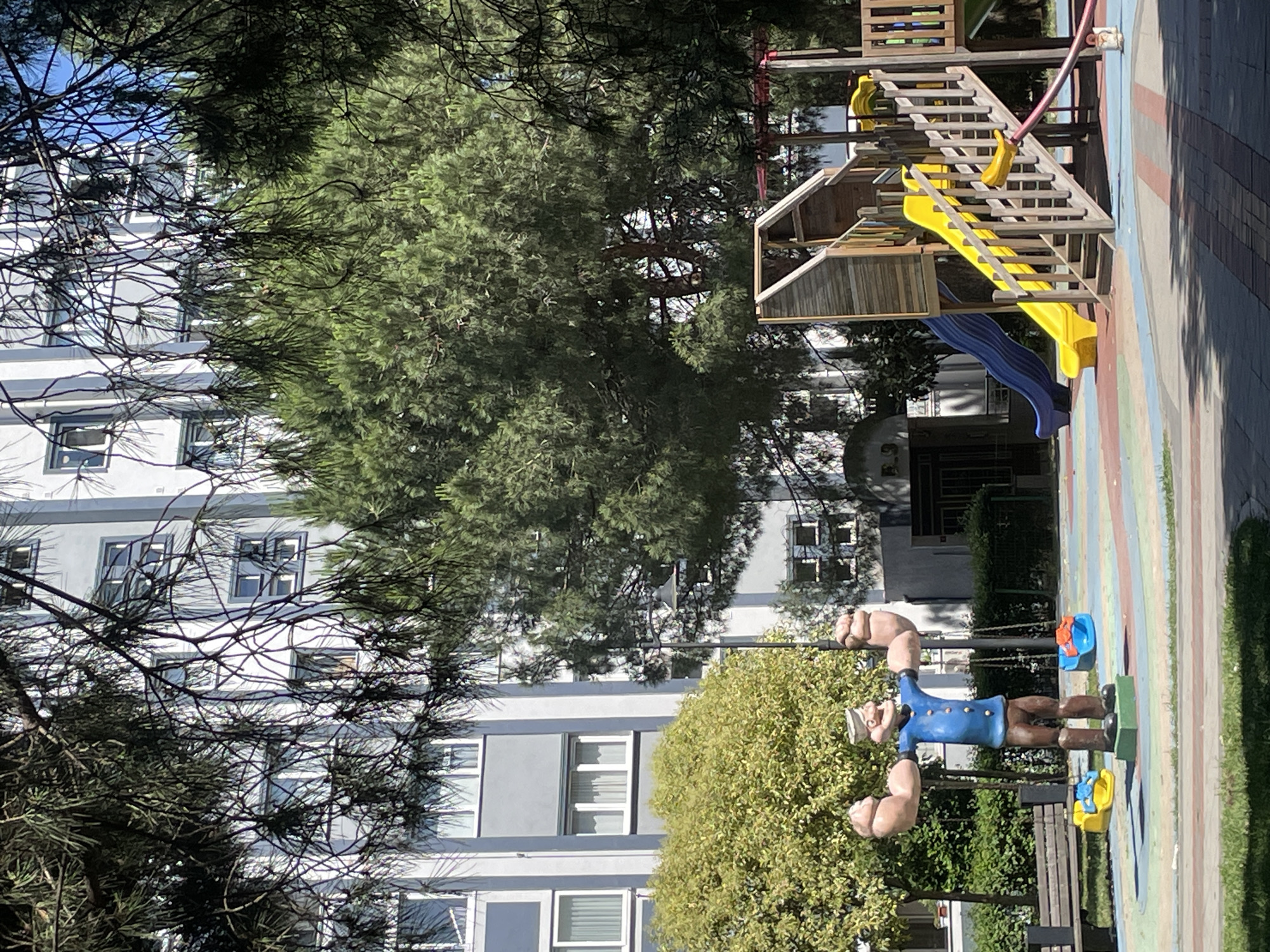 The image shows a cartoon statue of Popeye standing on a colorful playground. Popeye is depicted in his iconic blue shirt and white sailor hat, with his muscular arms raised. The playground features a wooden structure with slides, stairs, and a yellow ladder. There are also two small blue and yellow buckets visible on the ground near Popeye. In the background, a modern apartment building with many windows is partially obscured by trees. The scene appears to be outdoors on a sunny day, with shadows cast on the ground. The playground and the apartment building suggest a residential area. The overall impression is a cheerful and playful scene, likely in a park or public space. No visible text is present in the image.