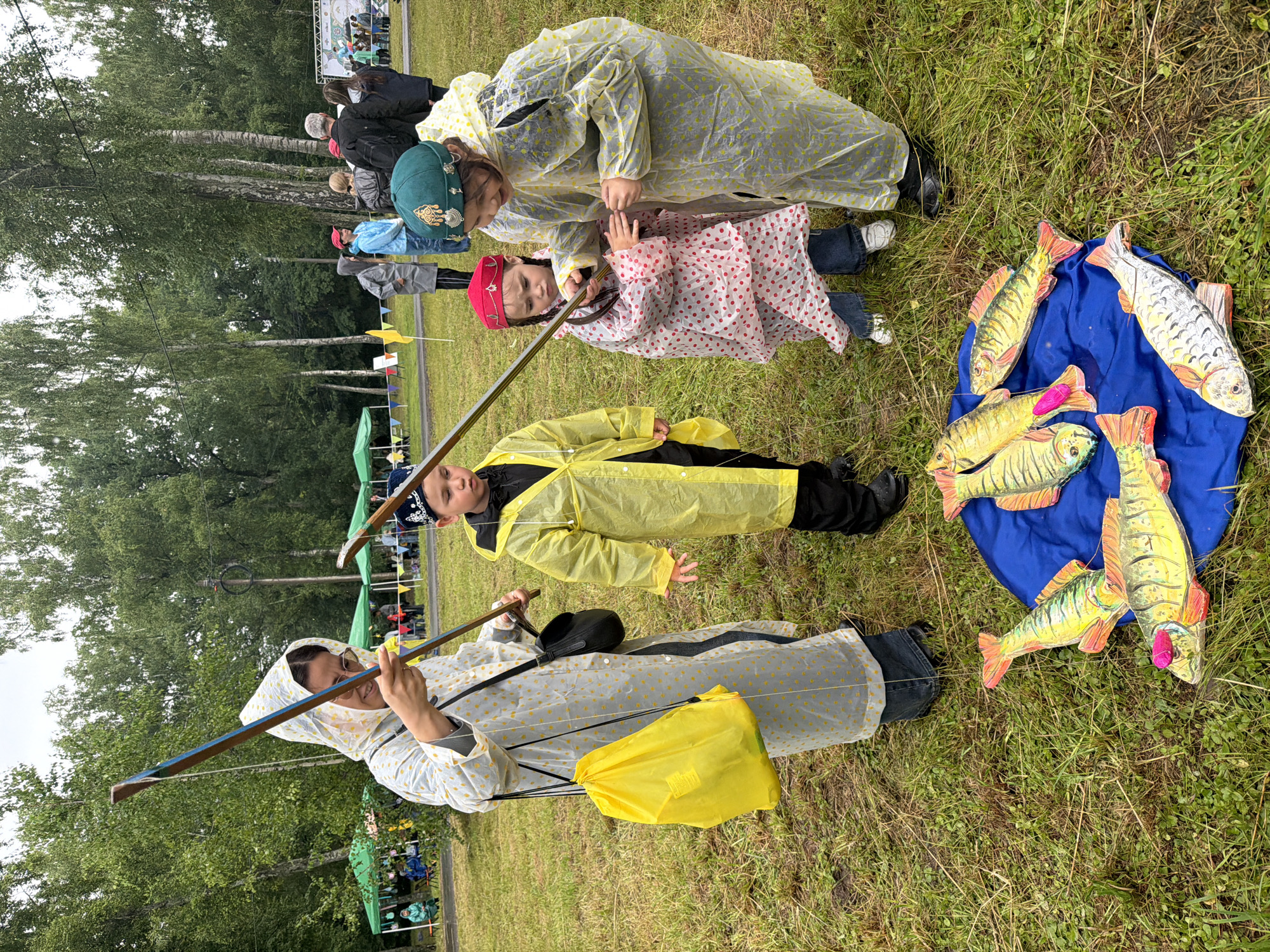 In an outdoor grassy area, likely a park or fairground, a group of people are gathered, with a focus on a few children dressed in raincoats and traditional headwear, interacting with fishing rods. The scene is set against a backdrop of tall trees and a cloudy sky, suggesting damp weather.
In the foreground, several colorful, stylized wooden fish are laid out on a blue tarp. The fish appear to be decorative or prizes.
To the left, an adult woman in a light-colored, polka-dotted raincoat and a grey patterned hood is holding a fishing rod, looking down at the children. She has glasses and is carrying a bright yellow drawstring bag over her shoulder.
To her right, a young boy in a bright yellow raincoat and a dark beanie with a pattern is also holding a fishing rod, looking towards the fish.
In the center, a young girl in a pink and white polka-dotted raincoat, with a red and gold ornate headpiece, is also holding a fishing rod and looking intently at the fish.
To the right of the girl, another young girl, wearing a green cap with gold embroidery and a clear, patterned raincoat, is also holding a fishing rod and looking at the fish, with an expression of concentration or excitement.
In the middle ground, more people are visible, some also in rain gear, and tents are set up, possibly for an event or market. The overall impression is a community gathering, likely a festival or competition, in which the children are participating in a fishing-related activity. The presence of raincoats indicates the weather is either rainy or has recently rained.