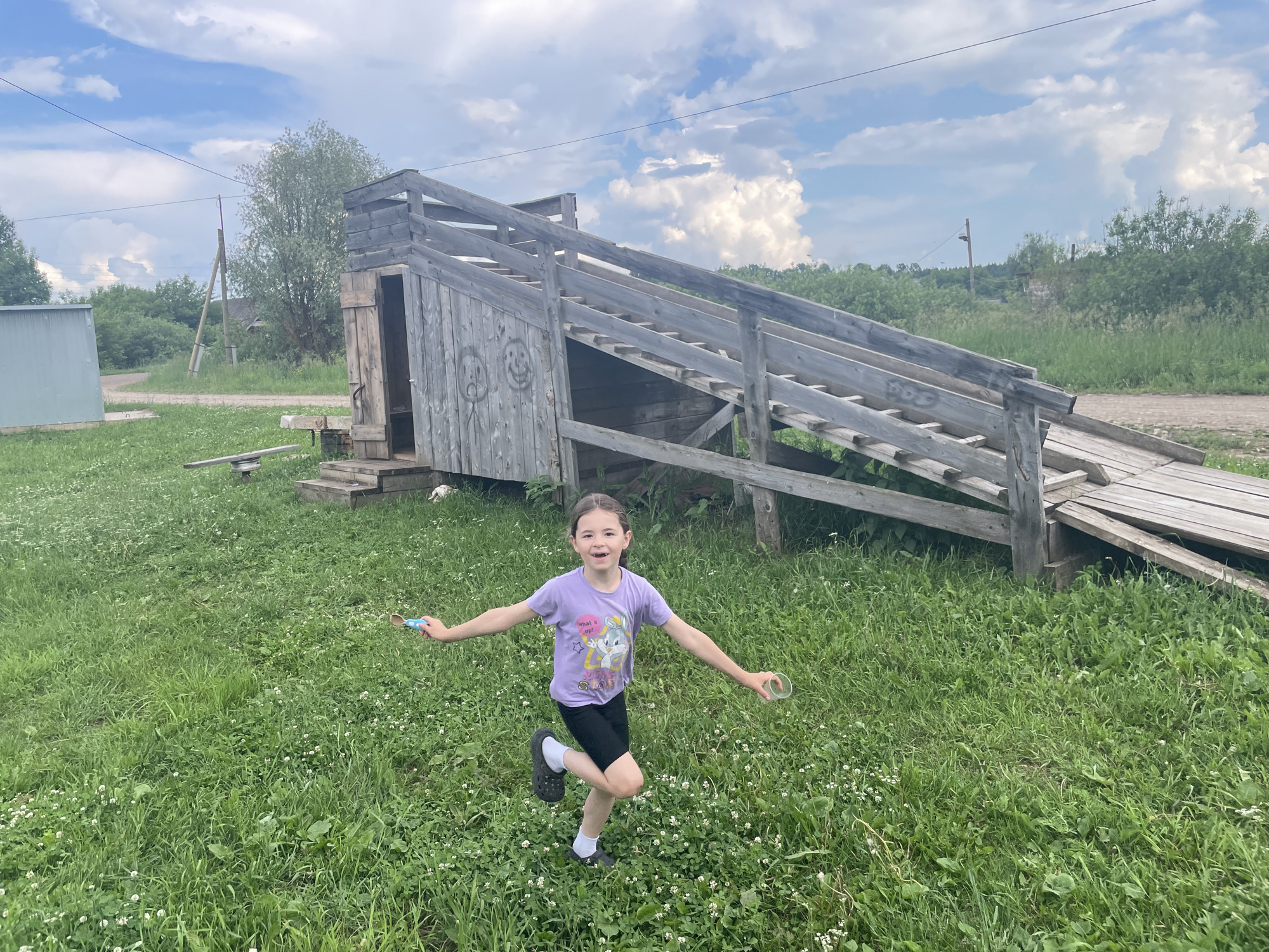 A young girl with brown hair, wearing a purple t-shirt and black shorts, runs across a grassy field. She has a joyful expression and is holding a blue object in her left hand and a clear plastic cup in her right hand. Behind her is a large wooden structure with a ramp, and in the background, there are trees and a cloudy sky. The scene appears to be in a rural setting in Nizhnii Bugrysh, Russia.