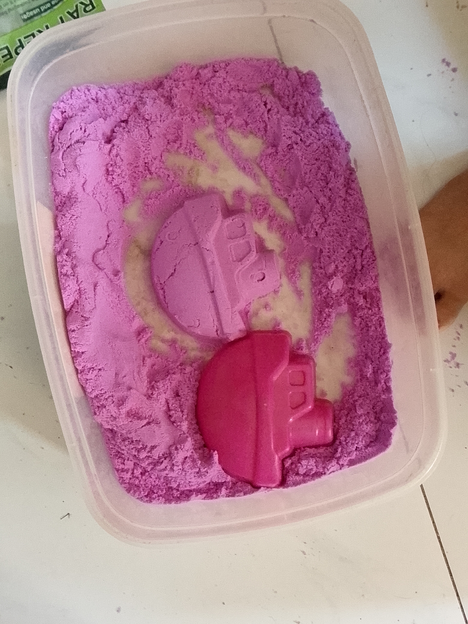 The image displays a clear, rectangular plastic container holding a quantity of vibrant purple kinetic sand. The sand exhibits a textured, granular appearance, with several impressions and disturbed areas, revealing lighter, almost beige patches beneath the surface.
Inside the container, two plastic molds shaped like castle or building components are present. One mold, in a lighter shade of purple or lavender, is largely submerged in the sand, leaving a circular impression around its edges. The second mold, a brighter hot pink, is partially embedded in the sand, with some purple grains clinging to its surface.
The scene suggests an ongoing or recently concluded play activity involving kinetic sand and molds. The container rests on a light-colored surface, likely a table or floor, which shows faint lines that could be tile seams. In the upper left corner, a section of a green and white box or package is visible, bearing partial text that reads "RATTLEPE". A small portion of a light-skinned human limb is visible on the right side of the frame, outside the container, and a few scattered specks of purple sand are also present on the white surface. The setting appears to be indoors with bright, even lighting.