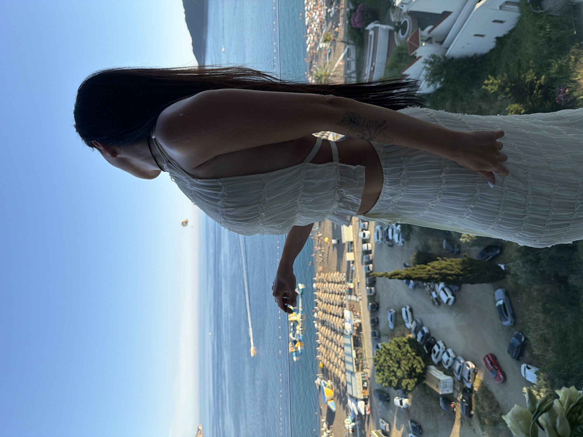 A woman stands on a balcony overlooking the city of Boreti, Montenegro. She is wearing a white two-piece outfit and has long, dark hair. Her head is tilted back, and she is looking up at the sky. In the background, the Adriatic Sea is visible, with several boats on the water. There is a beach with umbrellas and lounge chairs, and a parking lot with cars below. The sky is clear and blue. The image appears to have been taken in the late afternoon or early evening, as the sun is casting long shadows.