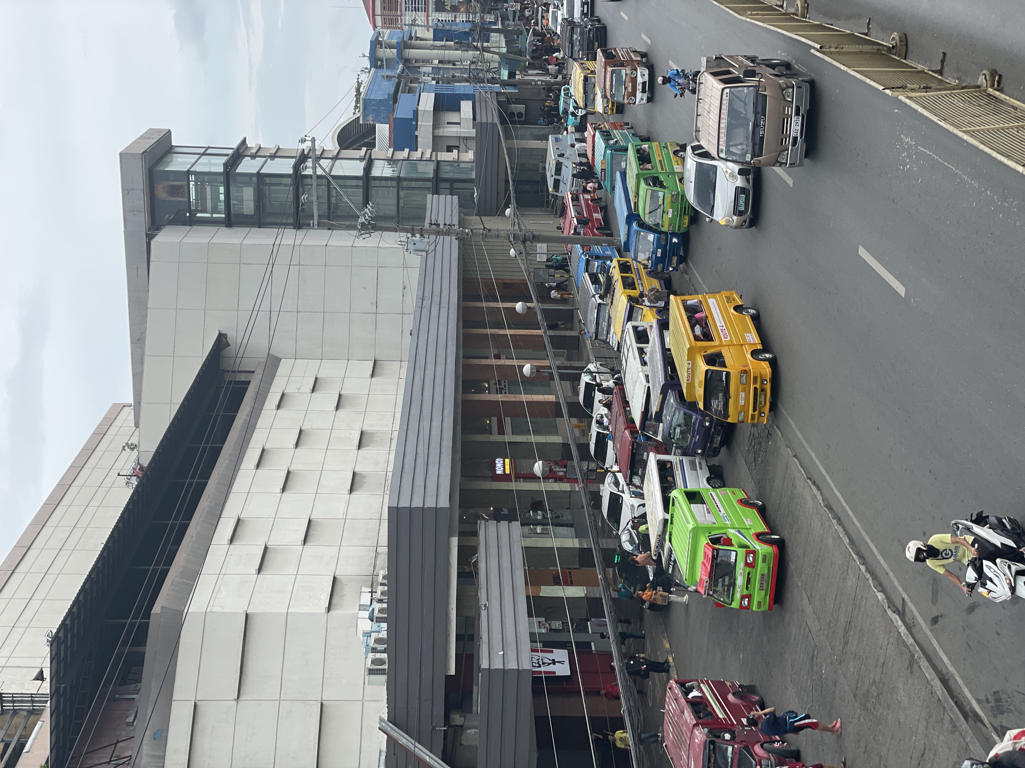 The image captures a busy street scene in Davao City, Philippines, with a focus on the proliferation of public utility vehicles, particularly jeepneys and vans, navigating a congested roadway. The architecture of the buildings lining the street suggests a commercial district, with a prominent modern building featuring extensive glass elements and a more traditional facade with repetitive paneling. A KFC restaurant is visible on one of the buildings. The sky is overcast, indicating a cloudy or potentially rainy day. People are seen walking on the sidewalks and some are inside the vehicles. The vehicles themselves are diverse in color and design, indicative of the varied public transport options in the city. A motorcycle rider in the foreground is also part of the traffic flow. The presence of numerous vehicles, pedestrian activity, and commercial establishments points to a lively urban environment within Davao City.