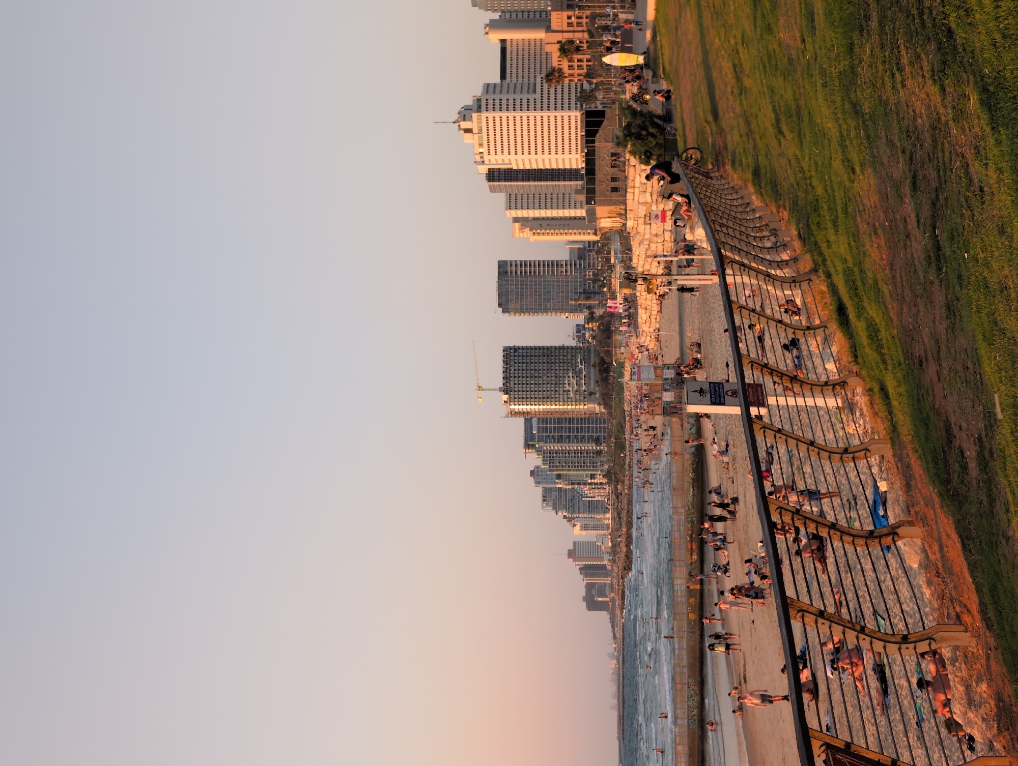 The image depicts a beach scene in Tel Aviv, Israel, at sunset. A wide expanse of sand is visible, populated by numerous individuals sunbathing, walking, and relaxing. The sea stretches out to the horizon, with gentle waves lapping the shore. A row of modern high-rise buildings lines the city's coastline, forming a distinct urban backdrop. A metal fence separates the beach from a grassy area in the foreground, where more people are seen enjoying the evening. The warm, golden light of the setting sun casts long shadows and bathes the scene in a serene glow.