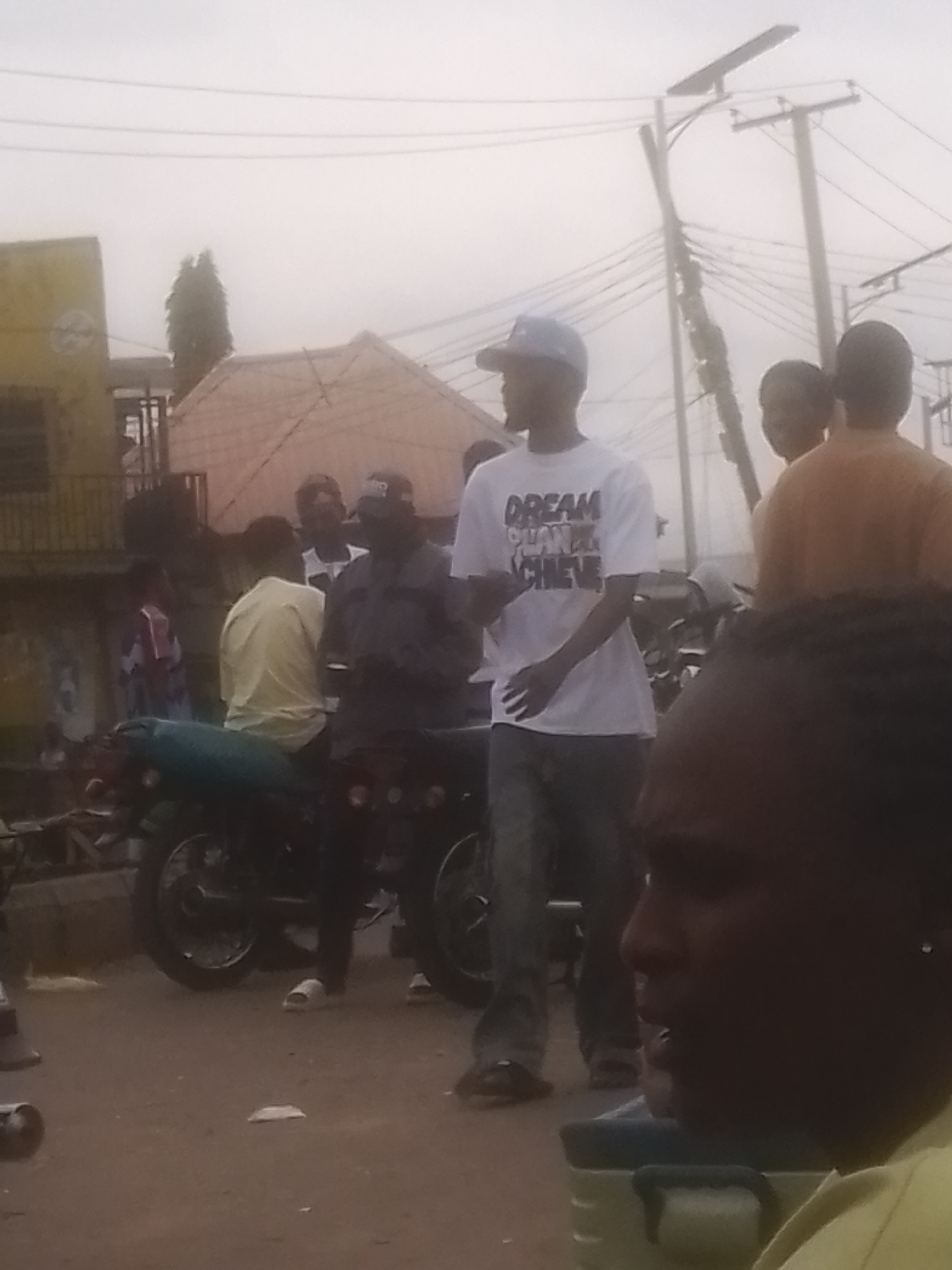 A medium shot captures a street scene with several individuals and a motorcycle. In the foreground, a person's face is visible in profile on the right side, partially obscured by a dark object. To the left, a person in a white t-shirt with the words "DREAM PLAN ACHIEVE" printed on it, wearing a baseball cap, stands facing right. Their left arm is extended. Behind them, another person in a yellow shirt is seated on a teal motorcycle. A person in a dark shirt and cap is also near the motorcycle. In the background, buildings and utility poles are visible under an overcast sky. The ground is a dirt or paved surface.