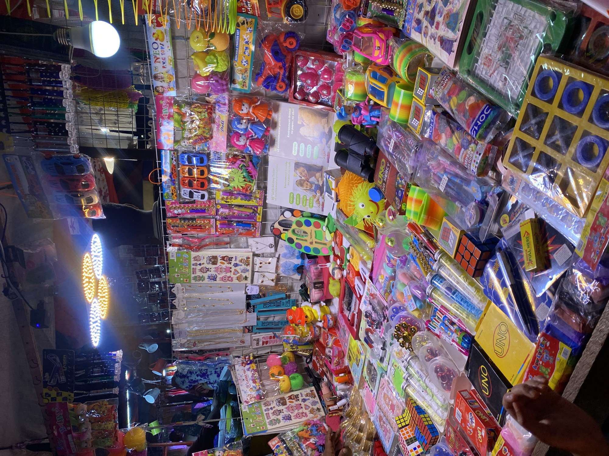 This is a medium-shot, eye-level photograph of a market stall displaying a variety of toys and novelty items, likely located in a busy urban area like Dhaka, Bangladesh. The stall is densely packed with merchandise, with items arranged on shelves and spread across tables.
In the foreground, a variety of colorful toys are visible. These include Rubik's cubes, UNO card games, small toy cars, and what appear to be plastic toys shaped like fruits. There are also some small dolls and various packaged stationery items. To the left, a person's hand is visible, suggesting interaction with the stall.
The middle ground reveals more toys and novelty items. There are several brightly colored plastic toys, including some that resemble bouncing balls and others that look like small animals. Packs of stickers featuring cartoon characters are also visible. Further back, there are shelves stocked with more items, including what appear to be small umbrellas and other packaged goods.
In the background, a ceiling with visible lighting fixtures can be seen. Several bright LED lights are illuminating the stall. The overall environment appears to be an indoor market or a covered street stall, with metal shelving and various hanging displays. The lighting is intense, creating strong highlights and some shadows, contributing to a bustling and vibrant atmosphere. The merchandise is primarily packaged, with a focus on bright colors and child-friendly designs.