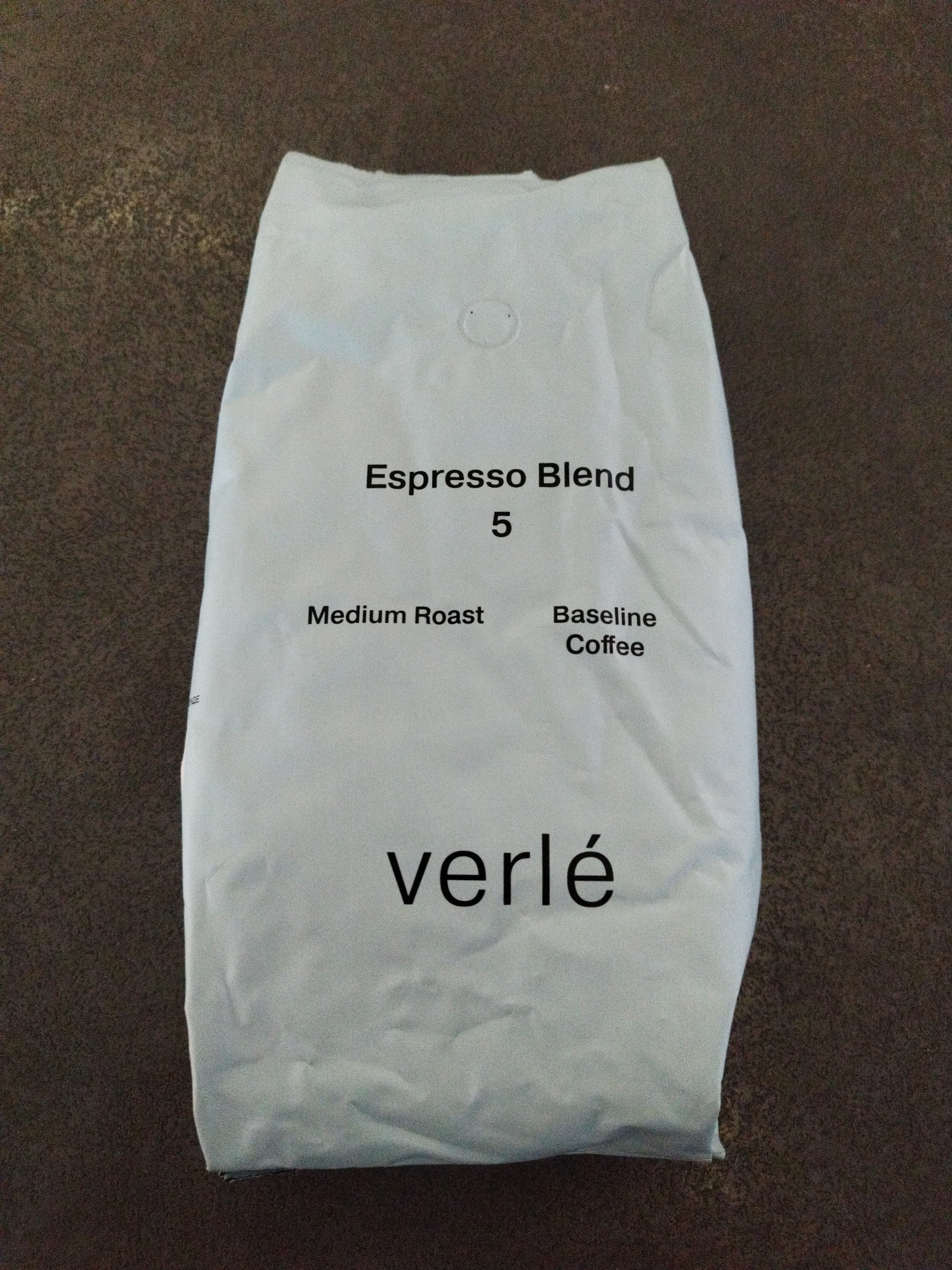 A white coffee bag is pictured against a dark brown, textured surface. The coffee bag has black text printed on it, indicating "Espresso Blend 5," "Medium Roast," and "Baseline Coffee." The brand name "verlé" is prominently displayed at the bottom of the bag in large, black lettering. There are no people, animals, or vehicles present in the image. The image does not provide any information about the location in the city of Saint Petersburg, Russia.