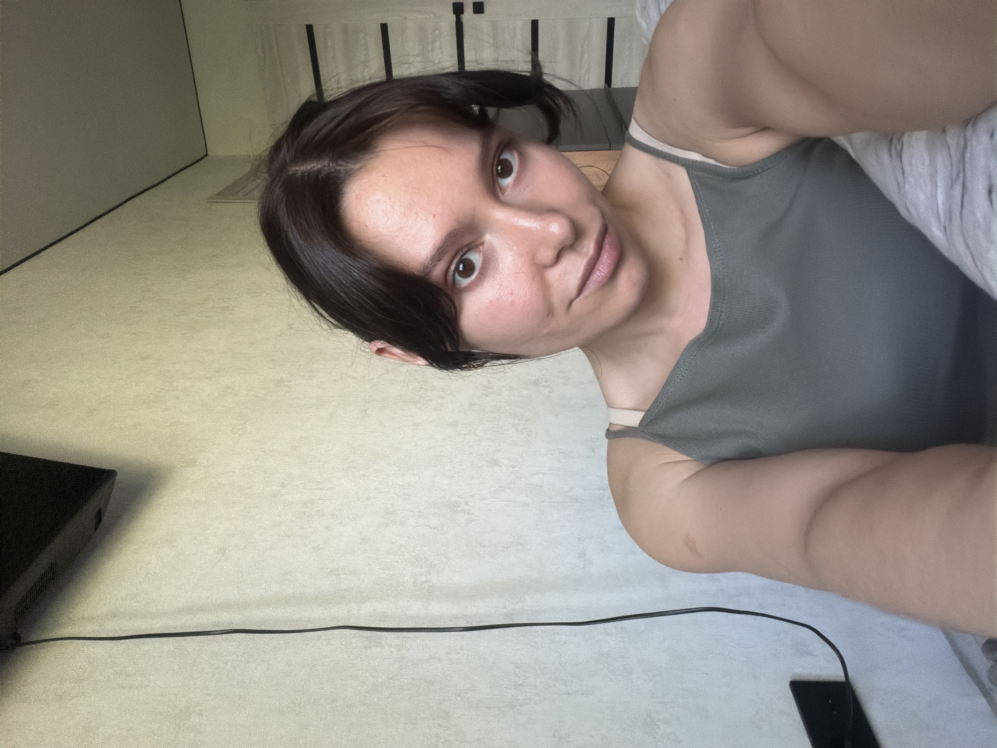 The image is a selfie taken indoors. A young woman with dark hair pulled back and some face-framing wisps is looking at the camera. She is wearing a grey tank top. Her skin appears natural, with some slight redness on her cheeks. Behind her, a textured wall with a black cable running down it is visible. A portion of a dark electronic device is mounted on the wall in the upper left corner. To the right, a doorway with black horizontal accents and a closed door is visible. The scene appears to be a residential interior. The location in the city of Chaikovsky, Russia cannot be determined from the visual information provided.