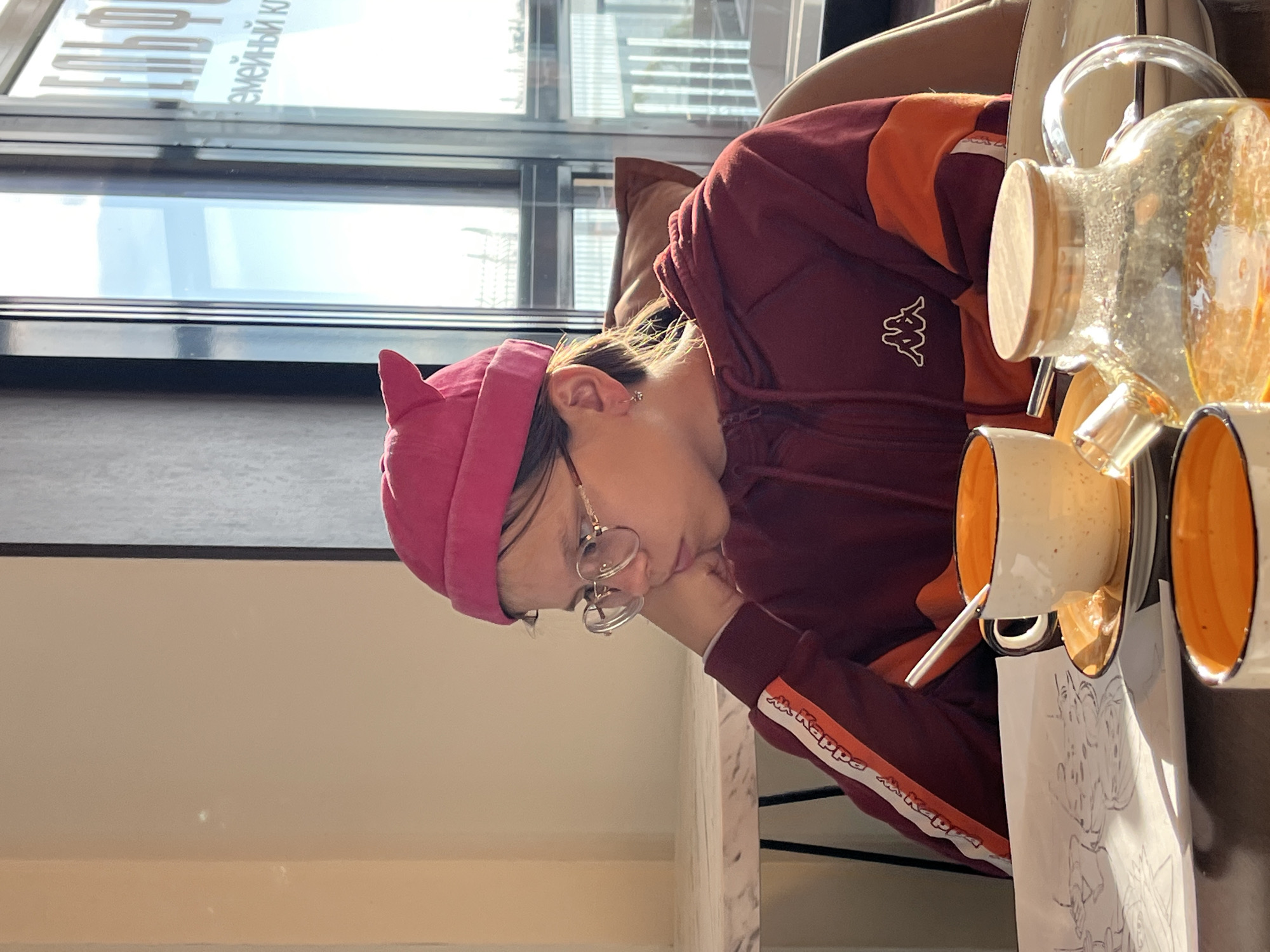 An indoor, well-lit scene captures a young person seated at a table, resting their head on their hand. The image is oriented vertically, showing the person in profile, facing towards the right side of the frame. She wears a unique pink beanie with small, pointed ear-like features, round gold-rimmed glasses, and a maroon and orange hooded sweatshirt with the white "Kappa" logo repeated on an orange stripe down the sleeve. An earring is visible in her left ear.
On the table in front of her, to the right, sits a clear glass carafe with a wooden stopper, filled with an amber liquid, possibly juice or tea. Below it are two white ceramic cups with vibrant orange interiors, and a white spoon is present in one of them. A partially visible sheet of paper with a black and white sketch, possibly a doodle or cartoon, lies beneath the cups.
The background features a large window that lets in ample natural light, indicating it is daytime. Through the window, reflections of Cyrillic text are visible, which appears to include "ЕМЕЙНЫЙ КУХНЯ" (Yemeynyy Kukhnya), suggesting "Family Kitchen" or a similar establishment, consistent with the location in Chaikovsky, Russia. The person's posture, with her head resting on her hand, conveys a sense of contemplation, rest, or possibly boredom.