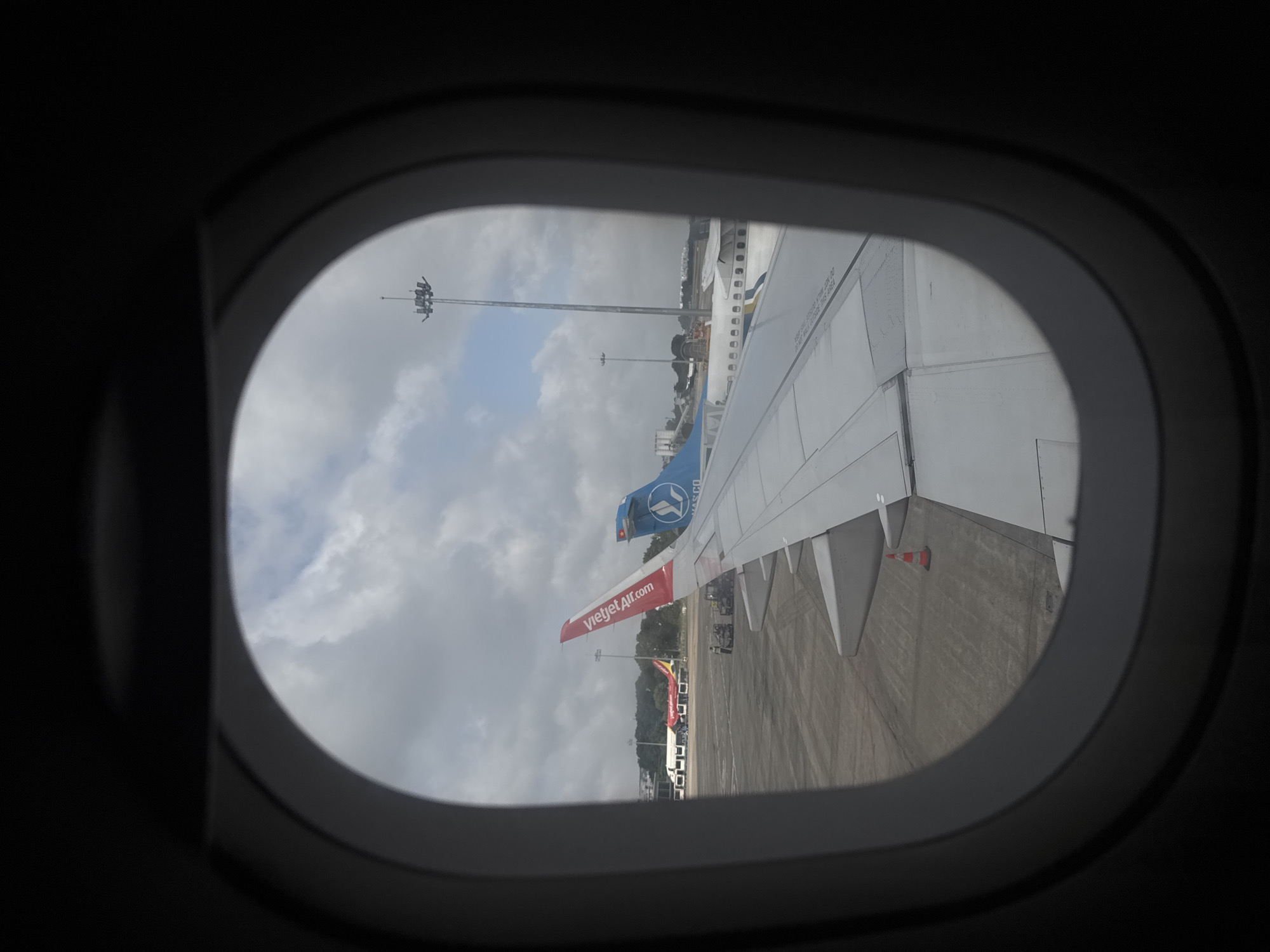 This image, taken from an airplane window, captures a tarmac scene at Tan Son Nhat International Airport, located in Thuận An, Vietnam. In the foreground, the wing of the aircraft the photographer is on is prominently visible. To the left, another aircraft with a red tail featuring the Vietjet Air logo is parked. Further to the right, a blue and white aircraft is visible, its tail featuring a logo with an avian design. Several other aircraft, buildings, and airport lighting towers are scattered across the tarmac under a cloudy sky. The ground is paved, with some vehicles and a traffic cone visible on the tarmac. A few individuals appear to be working on the tarmac near the Vietjet Air aircraft.