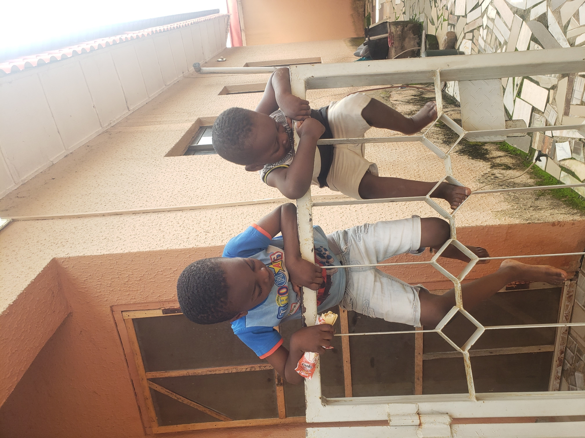 Two young boys are perched on a white metal gate in front of a two-story building in Sabon Tasha, Nigeria. The building's exterior walls are a rough, textured orange and light beige. A wooden framed window with a metal mesh is visible on the left side of the building. The ground is paved with irregular light-colored tiles and has some green moss growing in the cracks. A white gutter runs along the side of the building.
The boy on the left, wearing a blue t-shirt with red sleeves and light blue distressed denim shorts, is holding a wrapped snack in his left hand and appears to be eating it. His right hand is resting on the gate. The boy on the right, wearing a sleeveless striped top and light beige shorts, is leaning on the gate with his arms crossed and looking down. His body is partially obscured by the gate. The gate has a diamond-shaped pattern.