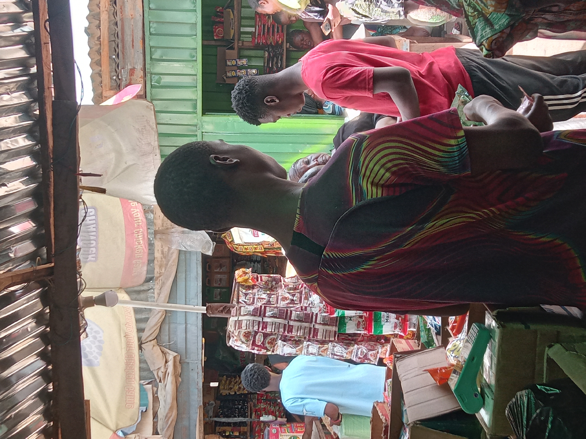 The image shows a street scene in Sabon Tasha, Nigeria. Several individuals are visible. One person wears a colorful patterned shirt. Another person in a red shirt is also seen. One individual is wearing a light blue shirt. A shop is in the background, with shelves stocked with various goods, including snacks and packaged items. Boxes are stacked near the shop entrance. The environment appears to be a bustling market area.
