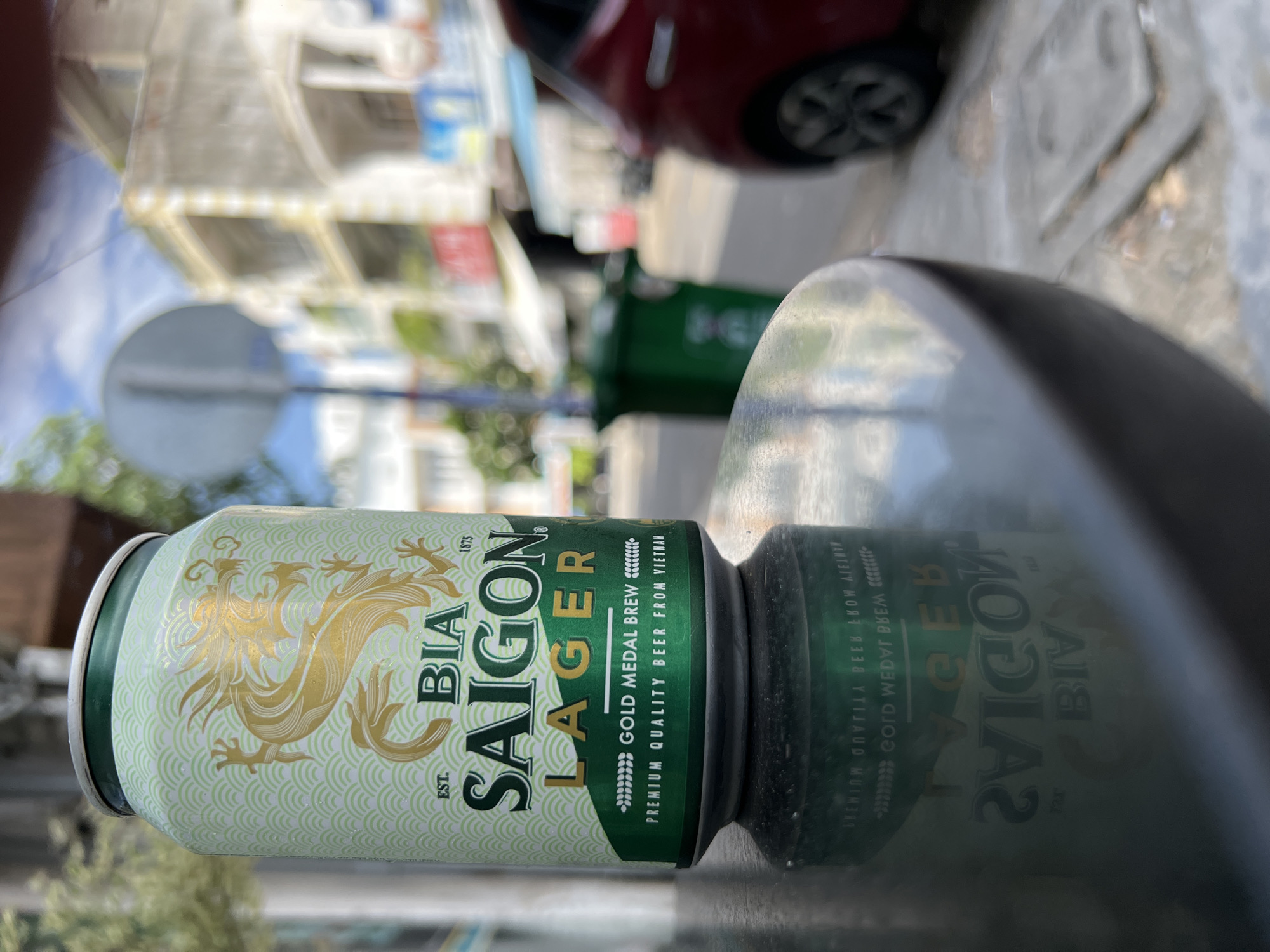 A close-up shot of a Bia Saigon Lager beer can is positioned on a reflective, dark grey surface in the foreground. The can features a light green background with a repeating wave pattern in a lighter shade, and a prominent golden dragon graphic on its upper half. The text "BIA SAIGON" is printed in large, bold, green lettering, with "LAGER" underneath in gold. Below this, smaller text reads "GOLD MEDAL BREW" and "PREMIUM QUALITY BEER FROM VIETNAM".
The background is blurred, suggesting an outdoor urban environment. A street scene is visible, with buildings with balconies, a green trash bin, a red car, and a circular street sign. The lighting suggests it is daytime, with a bright sky. The reflection of the can on the surface in the foreground shows reversed text and graphics, including "BIA SAIGON LAGER". The location is identified as Nha Trang, Vietnam.