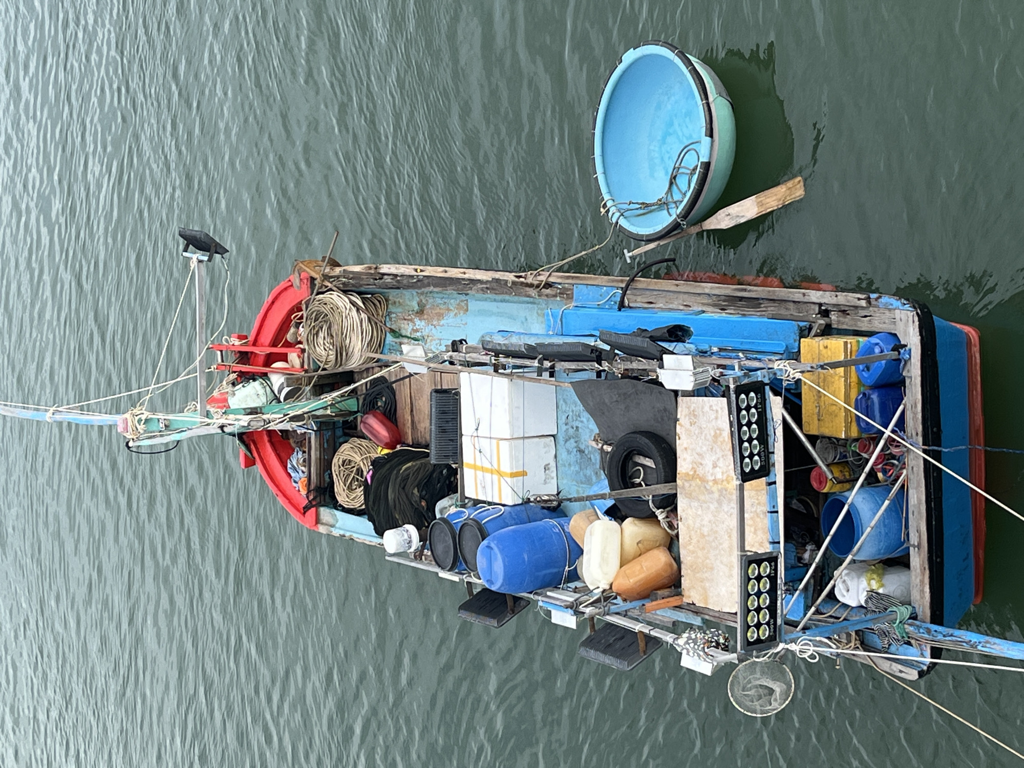 This is a high-angle, overhead shot of a loaded fishing boat docked in the waters of Nha Trang, Vietnam. The boat is elongated and appears to be made of wood, with a blue hull and a red-painted upper deck. The boat is filled with various supplies, including large blue barrels, white boxes, fishing nets, ropes, and yellow containers. Two floodlights are mounted on the side of the boat. A small, blue, circular basket boat is floating alongside the fishing boat, with a wooden oar resting inside it. The water surrounding the boat is a murky, greenish-blue. The sky is overcast, casting a dull light on the scene.